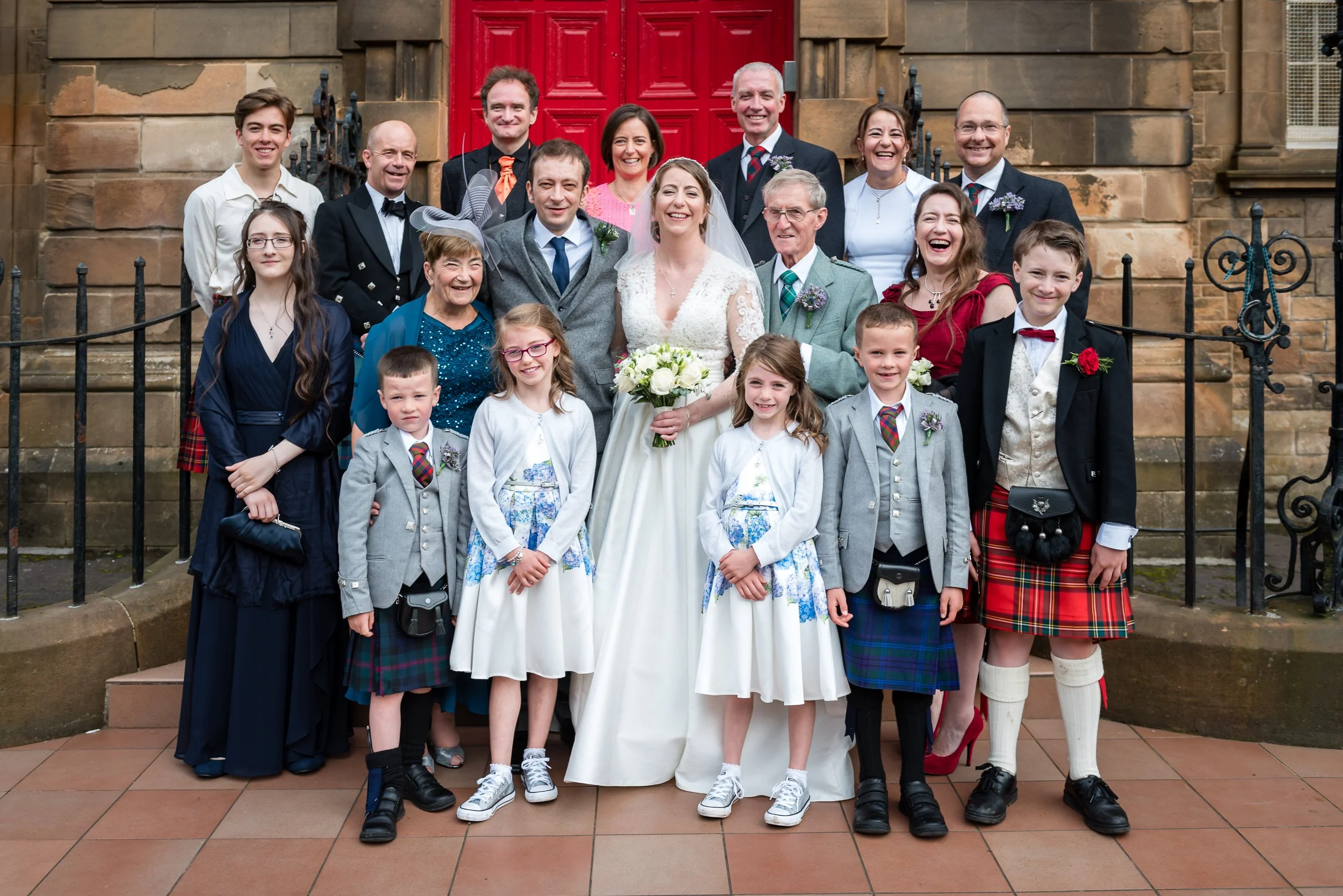 Group of people celebrating a wedding in front of a stone building with red door, including the bride in a white dress holding a bouquet, the groom in a gray suit, and family members dressed in formal, traditional, and Scottish attire.