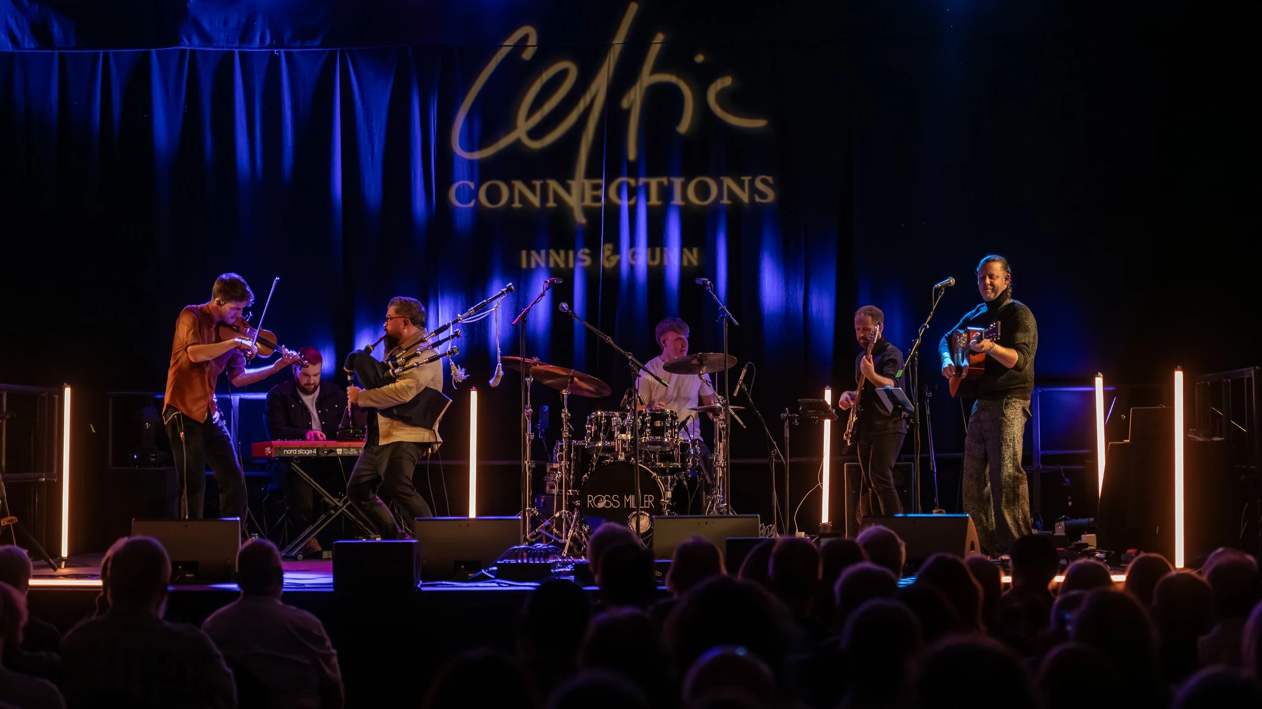 A band performs on stage, playing instruments, including bagpipes, a violin, keyboard, drums, bass, and guitar. The background features a blue curtain and a sign that says 'Celtic Connections Innis & Gunn.' The stage is lit with blue lighting.