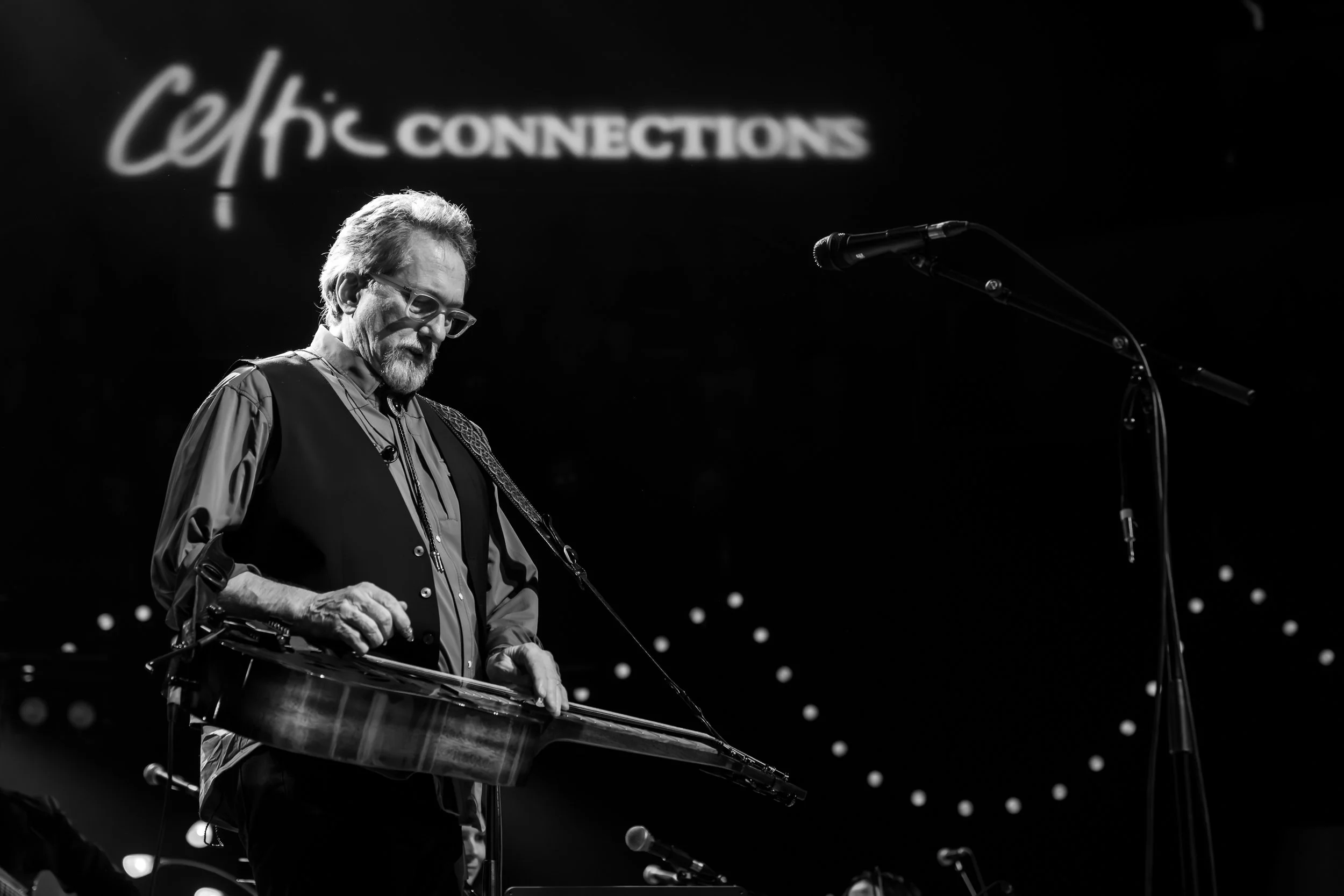 Black and white image of a man playing a steel guitar on stage with a microphone and a projected logo in the background. The logo reads, 'Celtic Connections'.