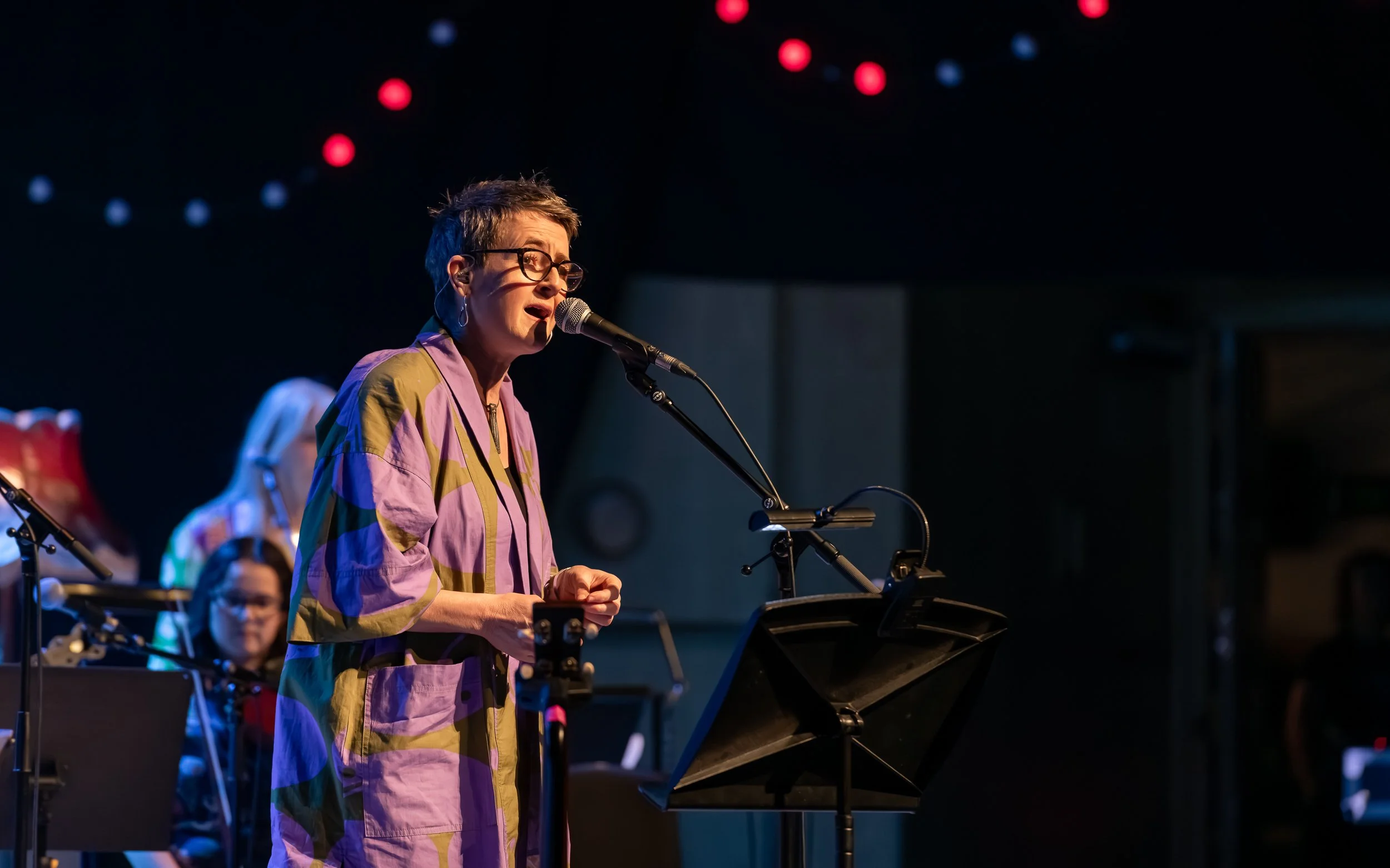 A woman with short hair, glasses, and earrings singing into a microphone on stage, with a music stand and musicians in the background, decorated with red and blue lights.