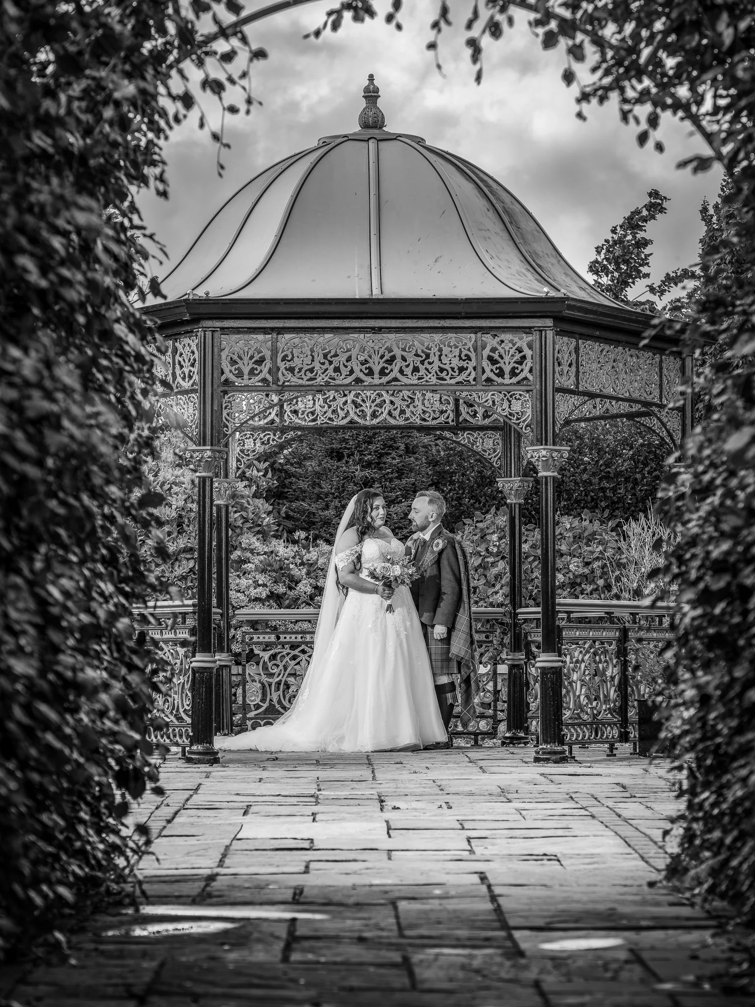 A bride and groom stand in an ornate gazebo outdoors, exchanging vows or sharing an intimate moment on their wedding day, with trees and cloudy sky in the background.