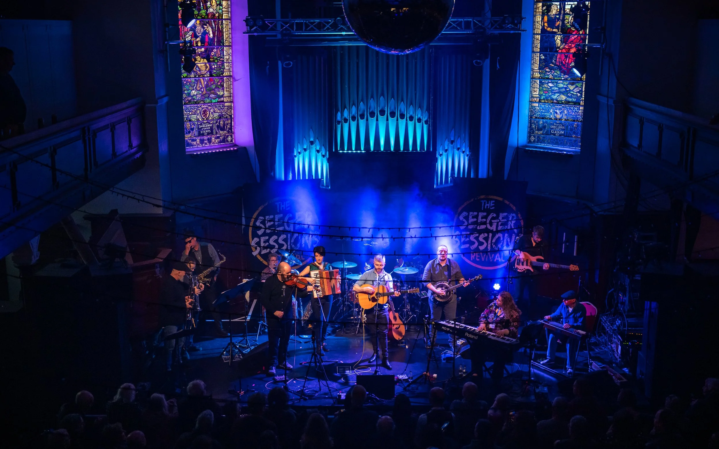 A band of multiple musicians performing on stage at a concert with blue lighting, a backdrop that reads 'The Seeger Session Revival,' musical instruments including guitars, violins, keyboards, and a bass, with stained glass windows and an audience vi