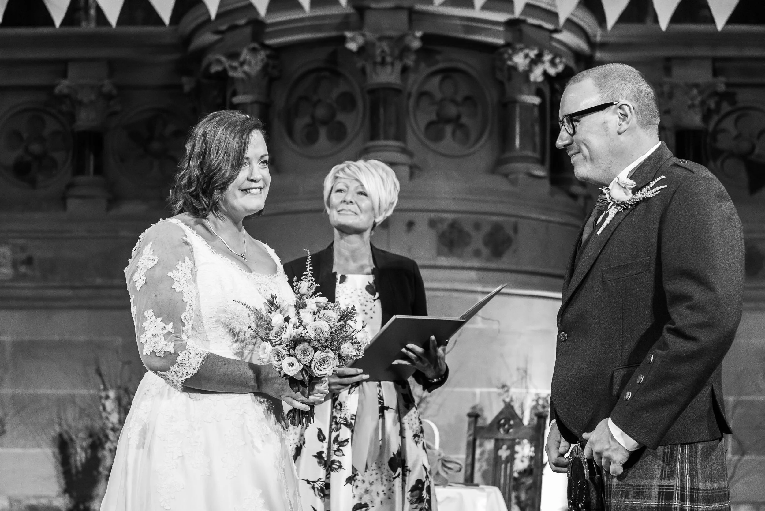 A black and white photo of a wedding ceremony inside a church, showing a bride in a lace dress holding a bouquet looking at the groom, who is dressed in a kilt suit, facing her. An officiant stands between them.