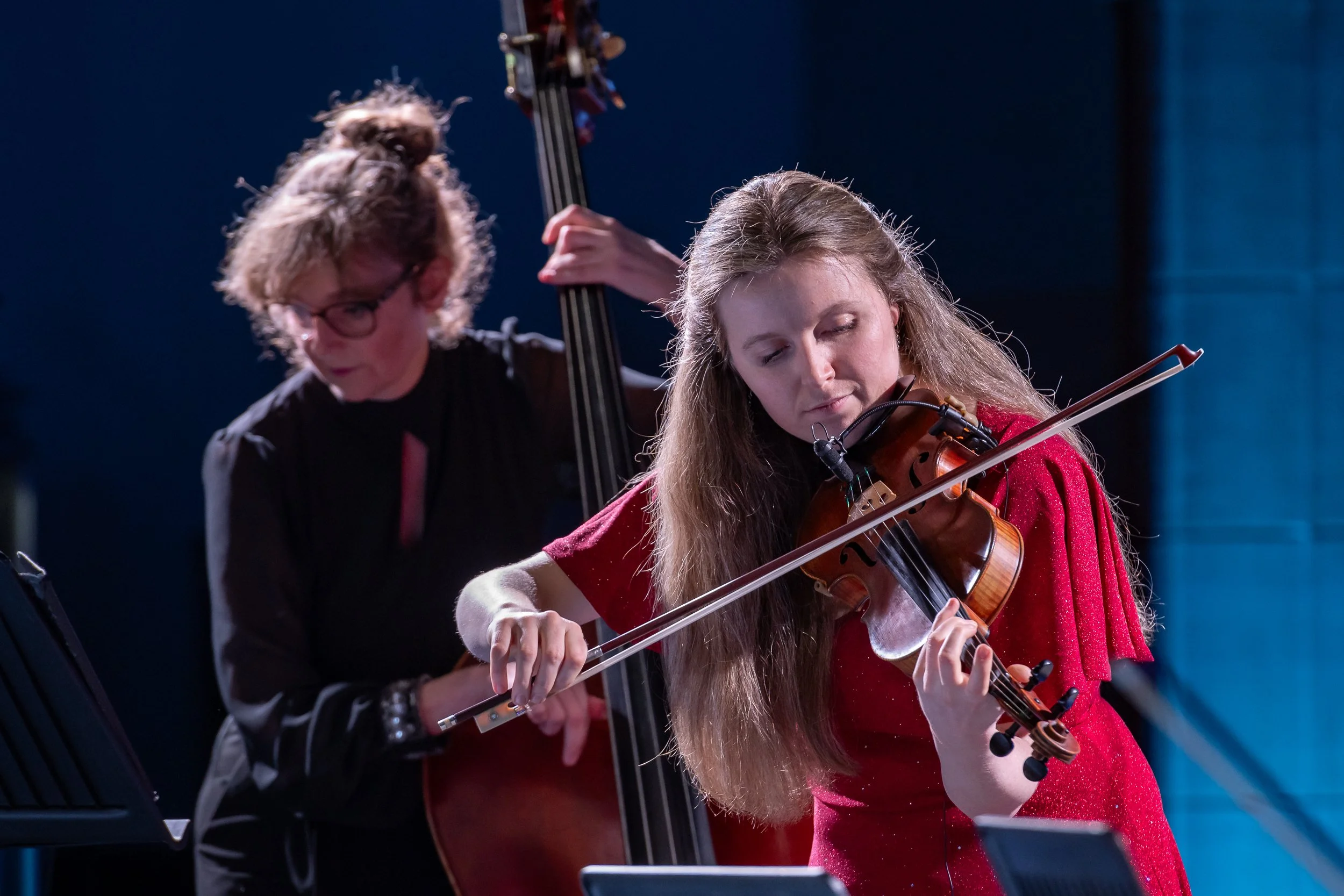 Two women playing musical instruments: one with a violin in the foreground, and another with a double bass in the background, performing on stage.
