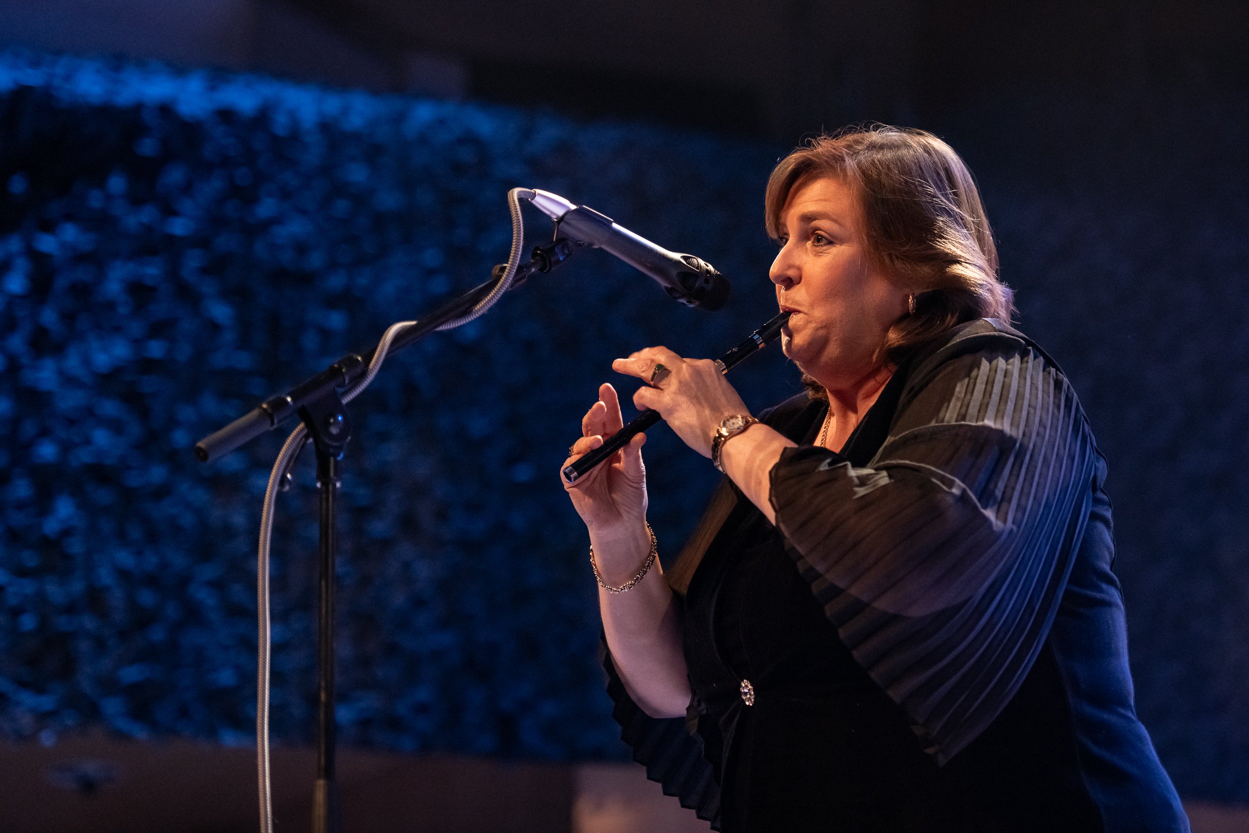 A woman with shoulder-length hair playing the whistle on stage with a blue textured background.