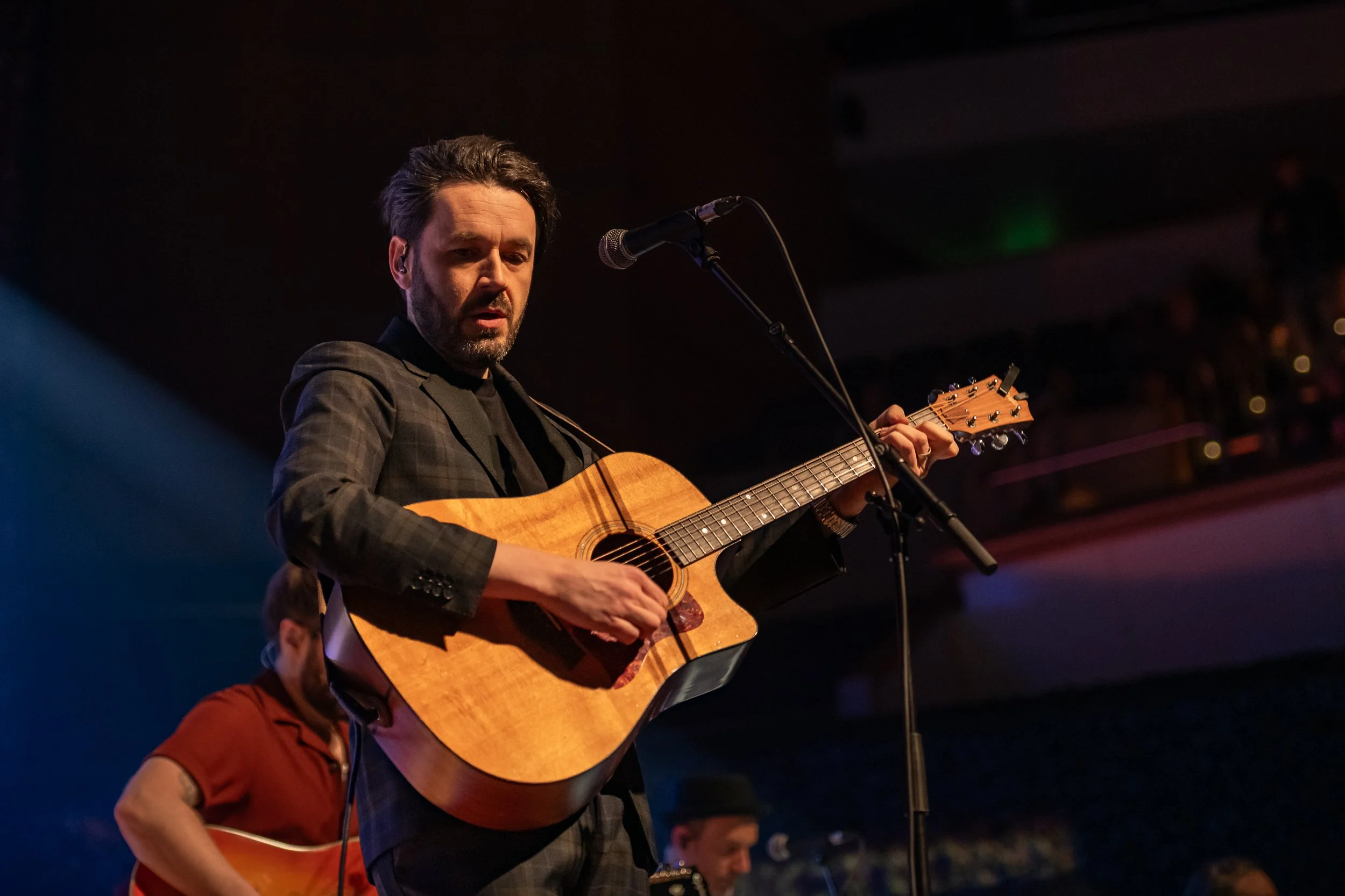 A man playing an acoustic guitar on stage during a live performance, with a smaller band member visible in the background.