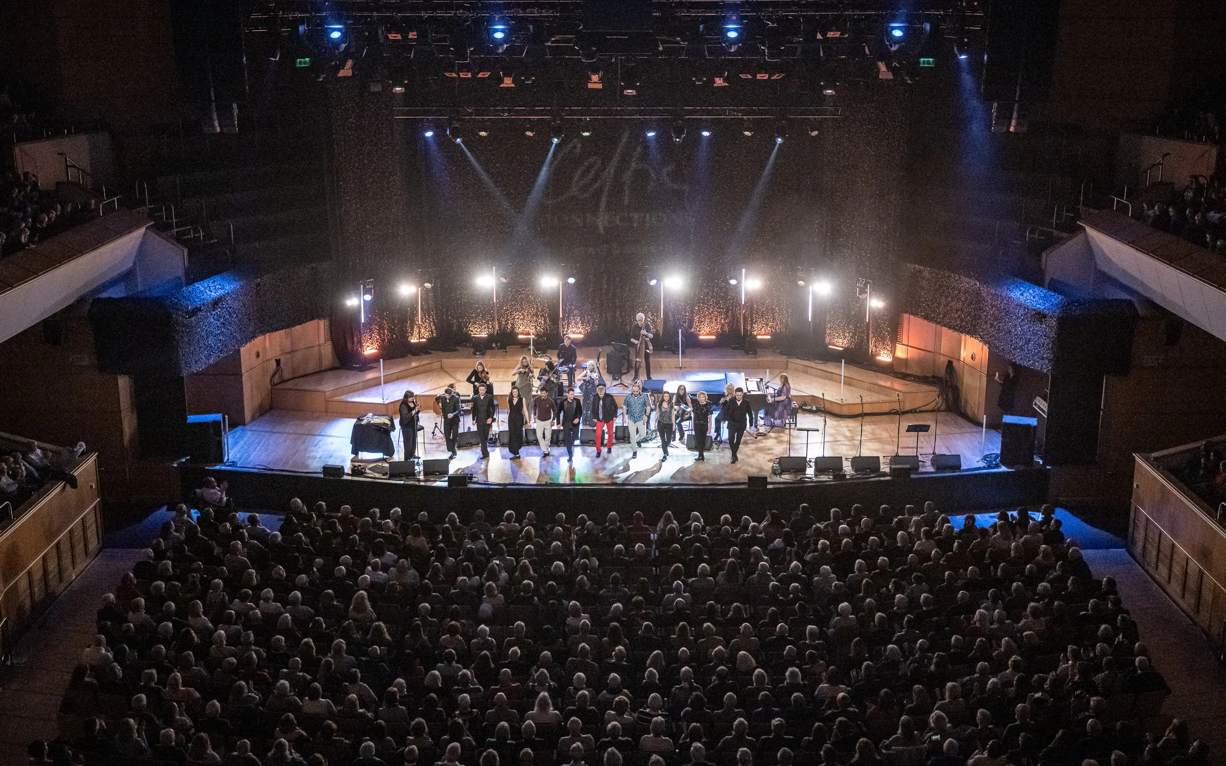 Concert hall filled with audience, performers on stage with musical instruments, stage with bright lights, and a dark backdrop.