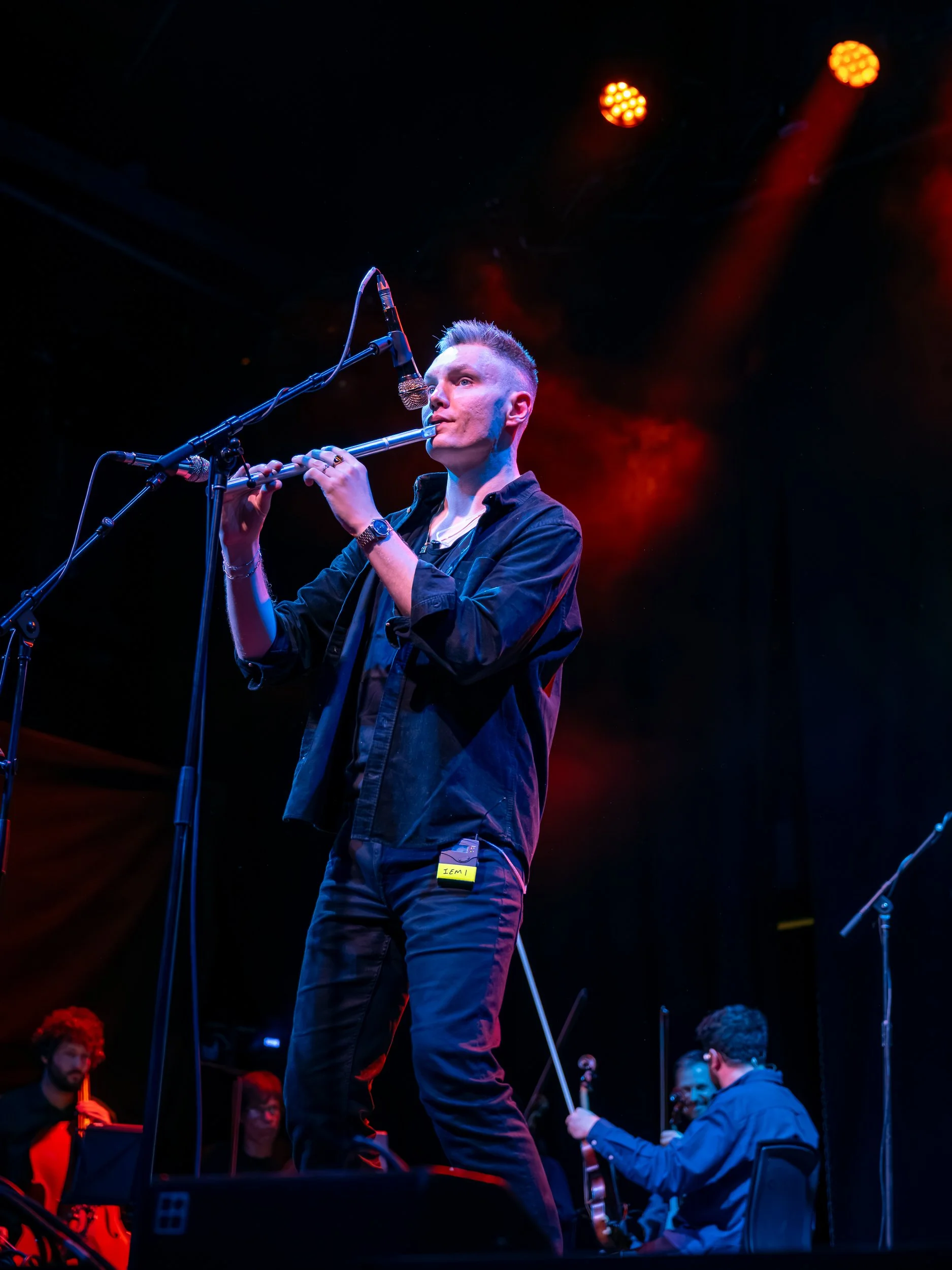 Musician playing a low whistle on stage with red and orange stage lights, and a string quartet in the background.
