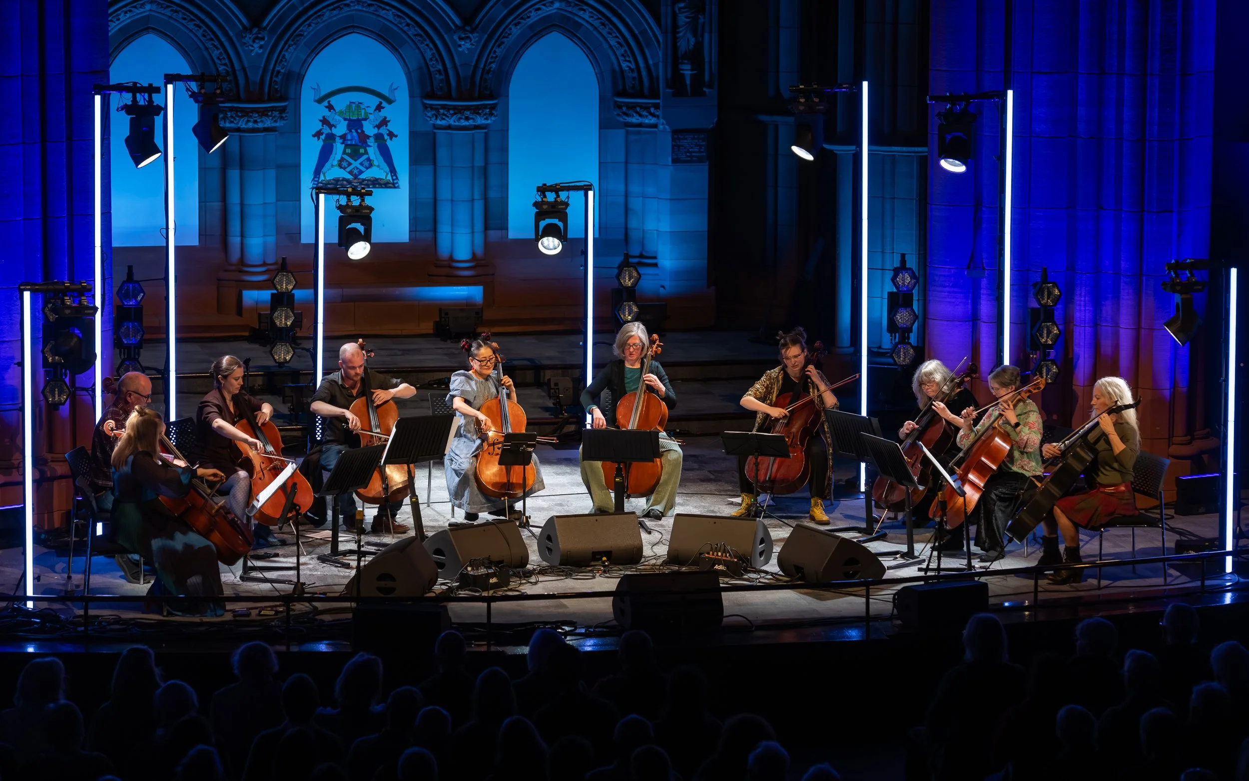 A cello orchestra performing on stage with blue lighting and neon light accents, audience visible in foreground.