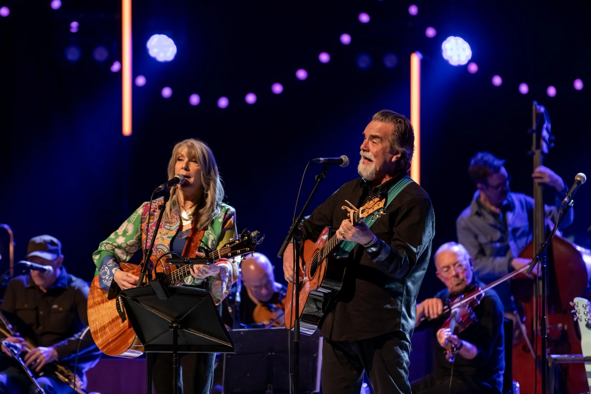 Two musicians singing and playing guitars on stage, with a band in the background and colorful stage lights.