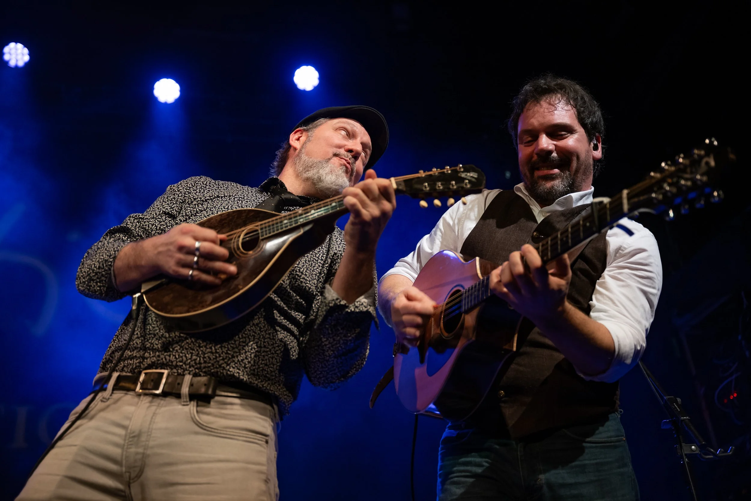 Two male musicians playing mandolin and bouzouki on stage with blue lighting in the background.