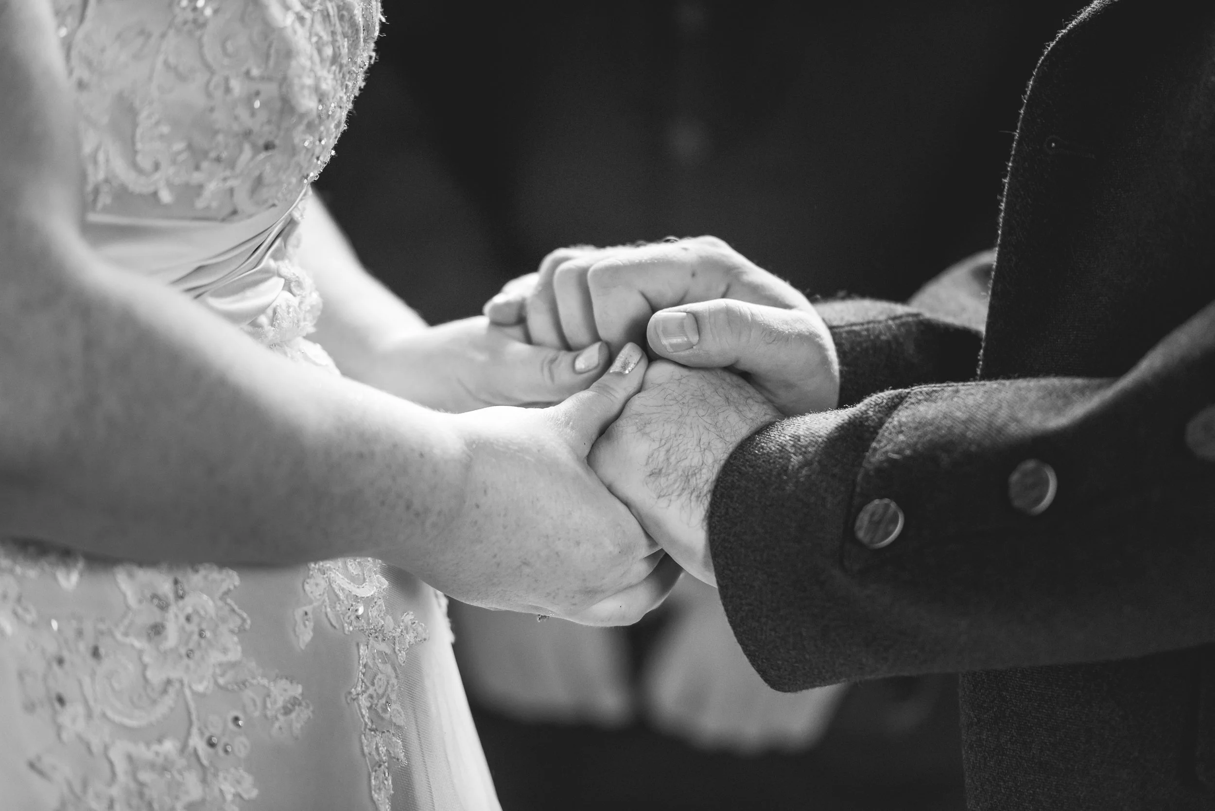 Close-up of a groom and bride holding hands during a wedding ceremony, with the bride wearing a lace wedding dress and the groom in a dark suit.