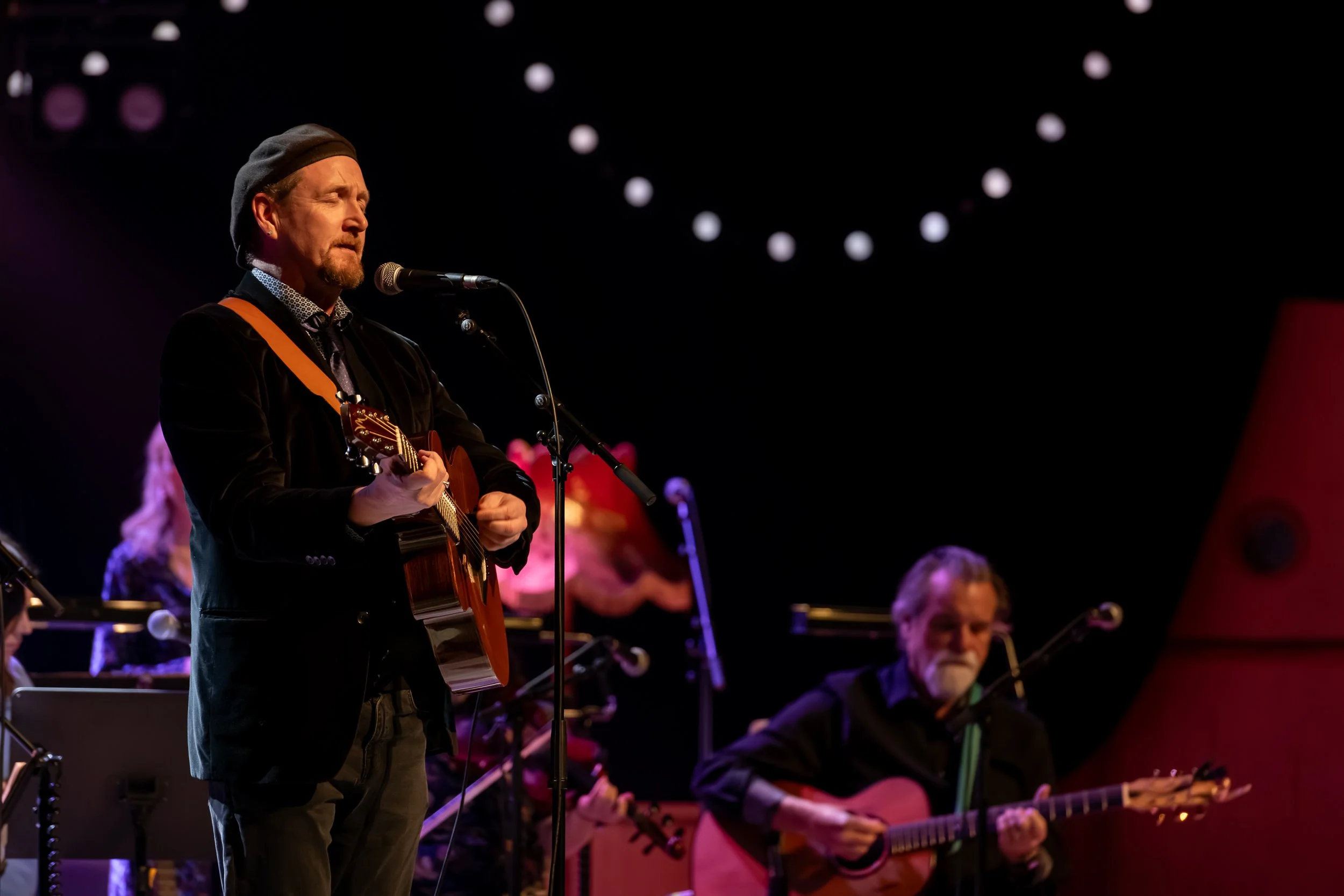 Musicians performing on stage with guitars, with one man singing into a microphone and others playing guitars, decorated with string lights in the background.