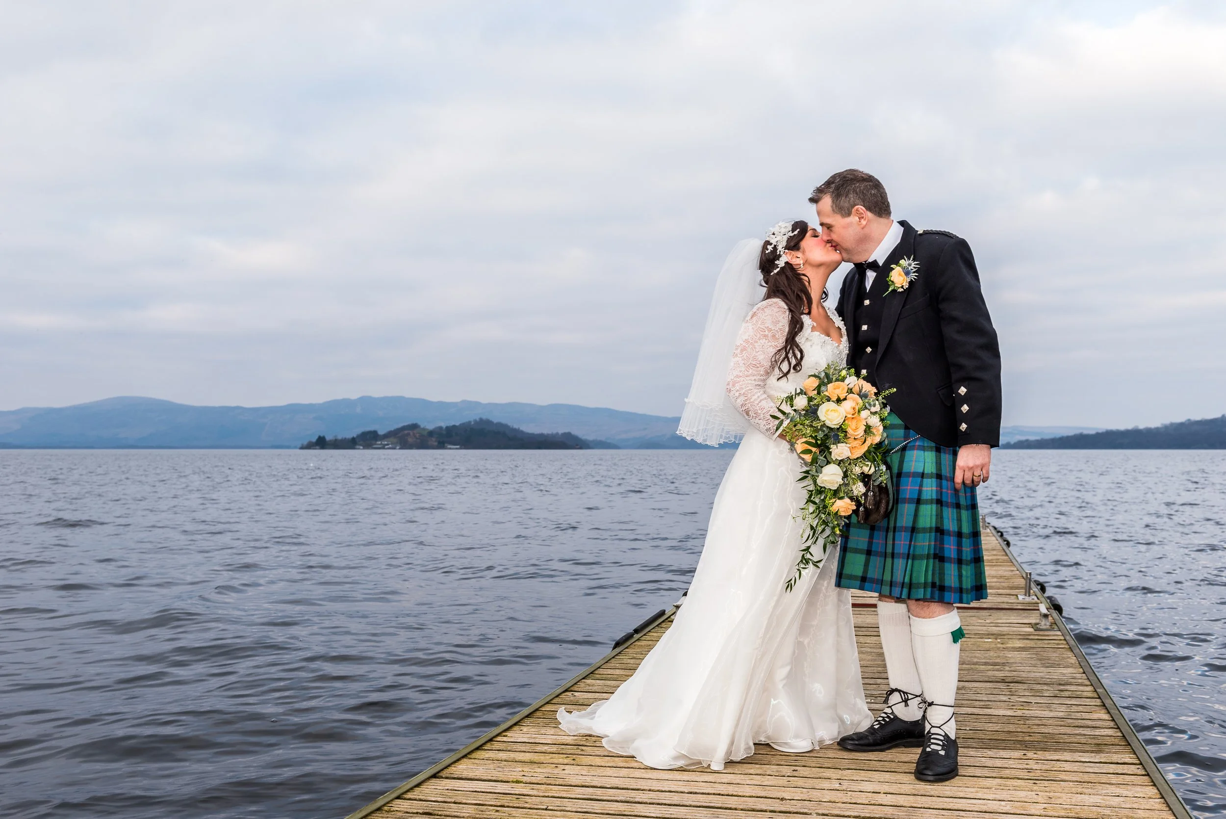Bride and groom kissing on a wooden dock by the water, with mountains and cloudy sky in the background. The bride wears a white wedding gown with lace sleeves, and the groom wears a traditional Scottish kilt with a black jacket, white shirt, and black shoes.