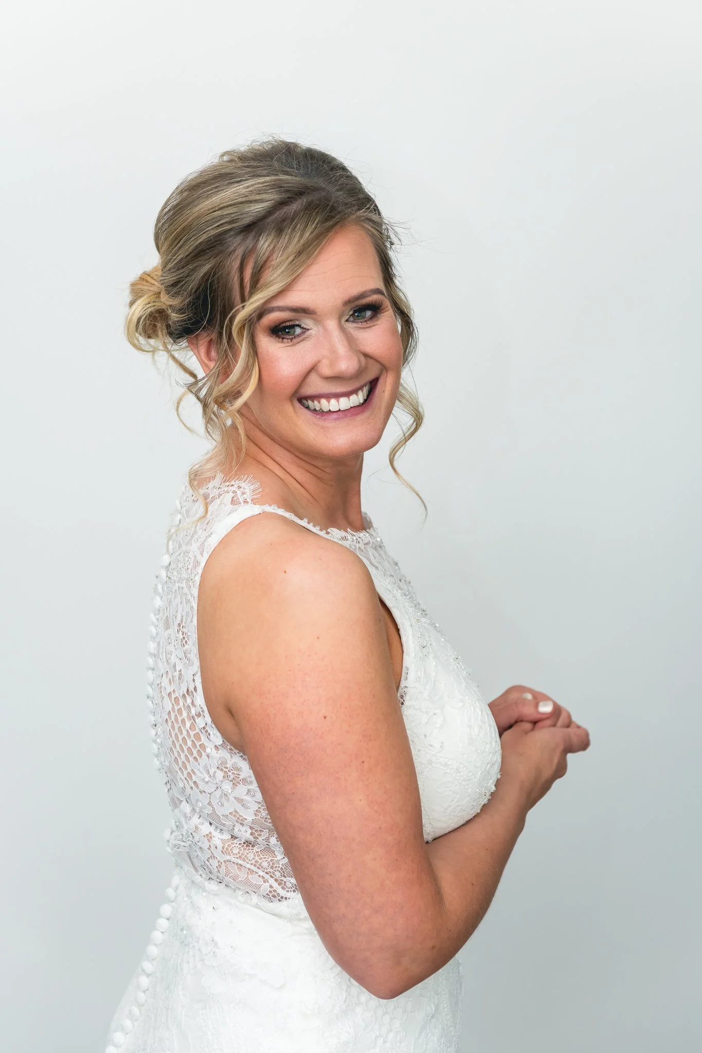 A smiling bride with blonde hair styled in loose curls wearing a white lace dress.