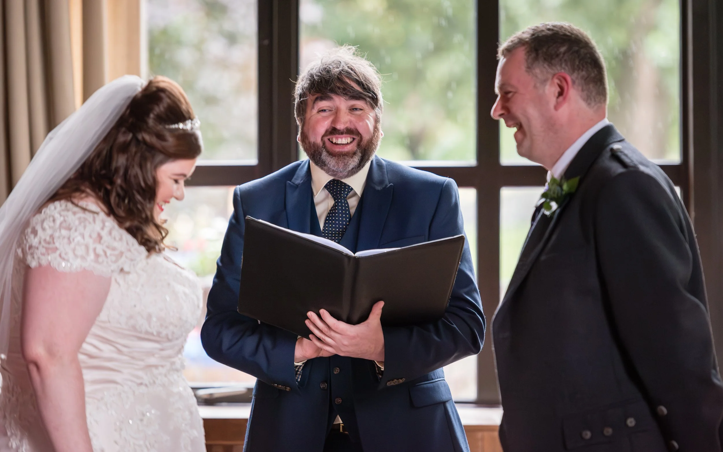 A bride and groom smiling during their wedding ceremony as the officiant reads from a book.