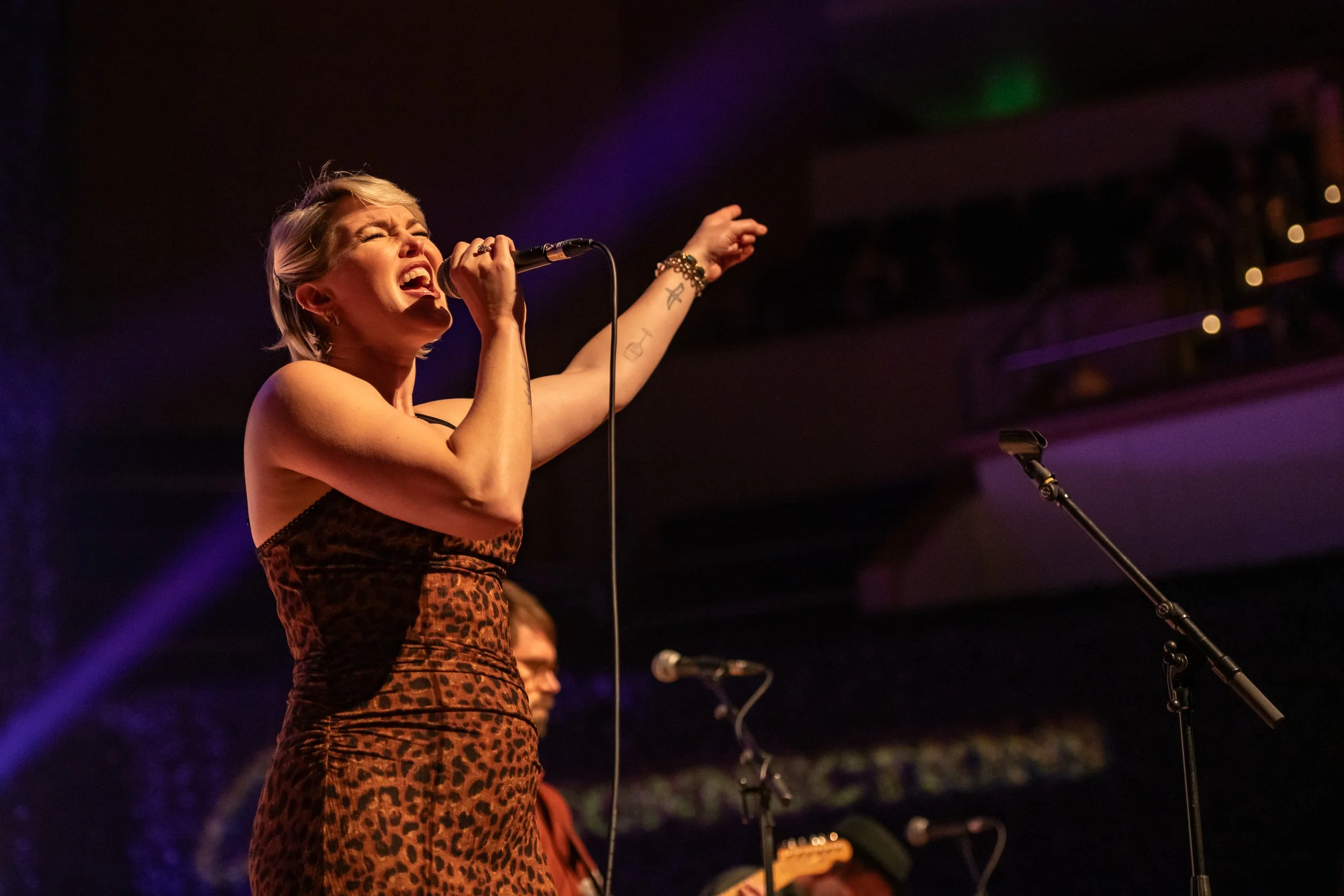 A woman singing passionately on stage, wearing a leopard print dress, with short blonde hair, holding a microphone in her right hand, with purple and green stage lights in the background.