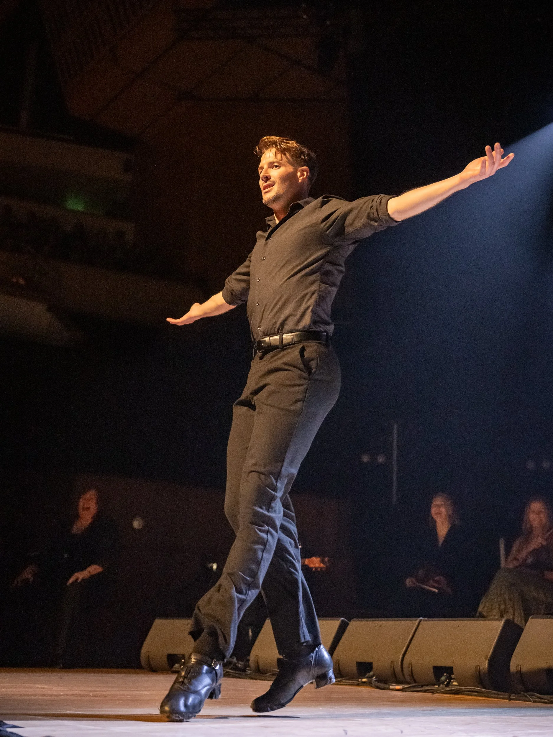 A man performing on stage with arms outstretched, wearing black pants, black shirt, and black dance shoes, with a dark background and band members watching.