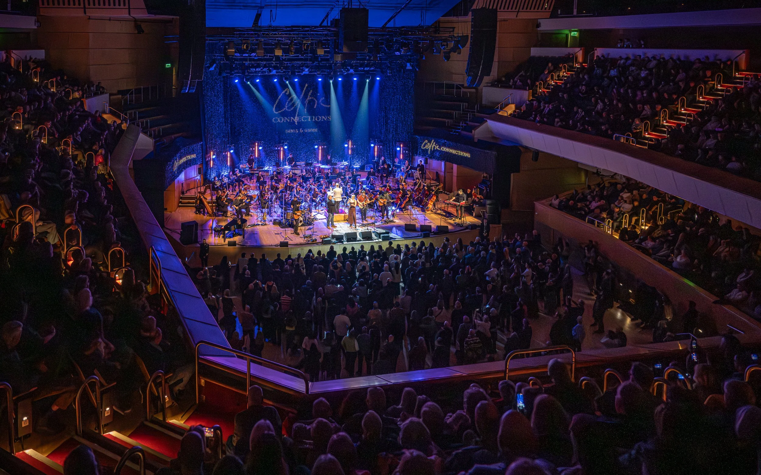 Concert hall with a large audience watching an orchestra perform on stage, illuminated by blue and purple lights.