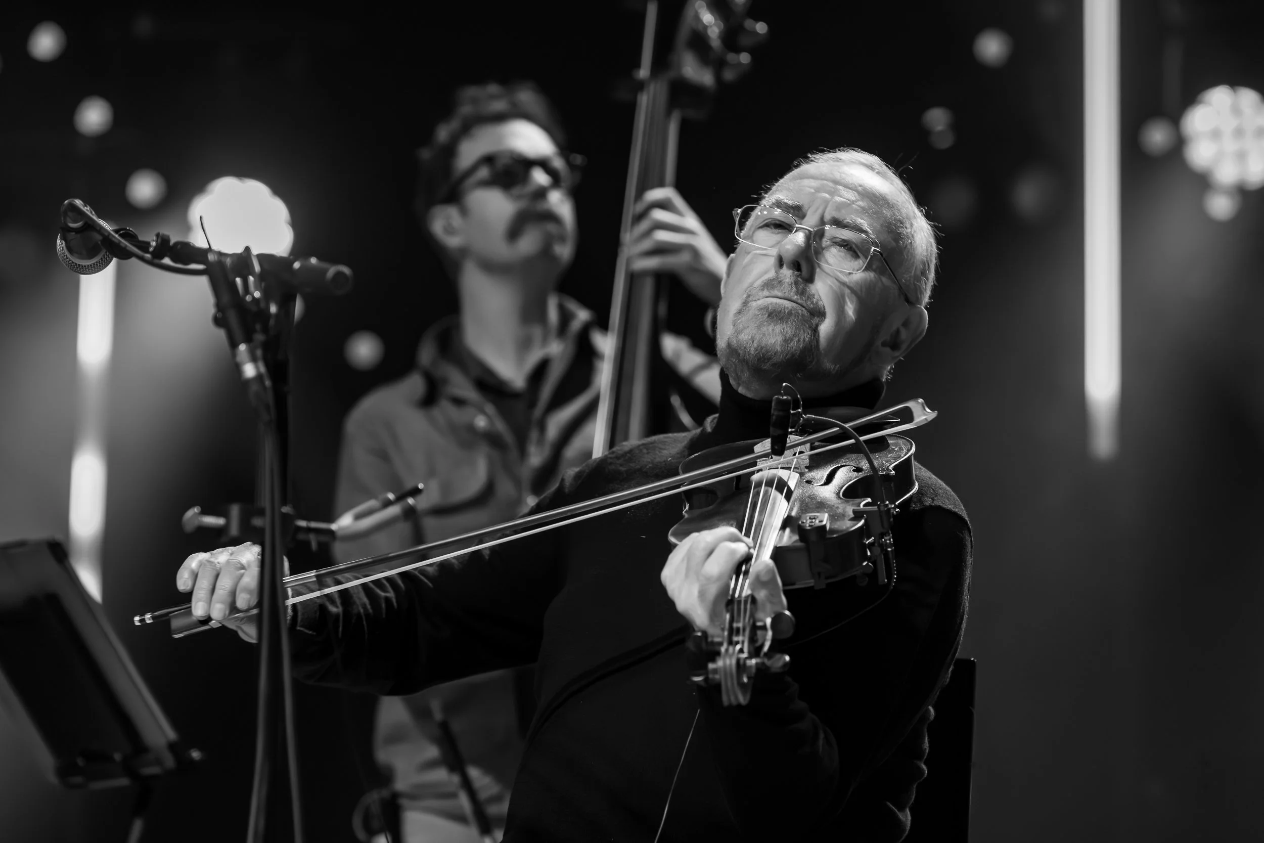 Two musicians playing string instruments on stage, one with glasses and a serious expression in the foreground, playing the violin, and another with a moustache and glasses in the background, playing double bass, in a black and white concert setting.