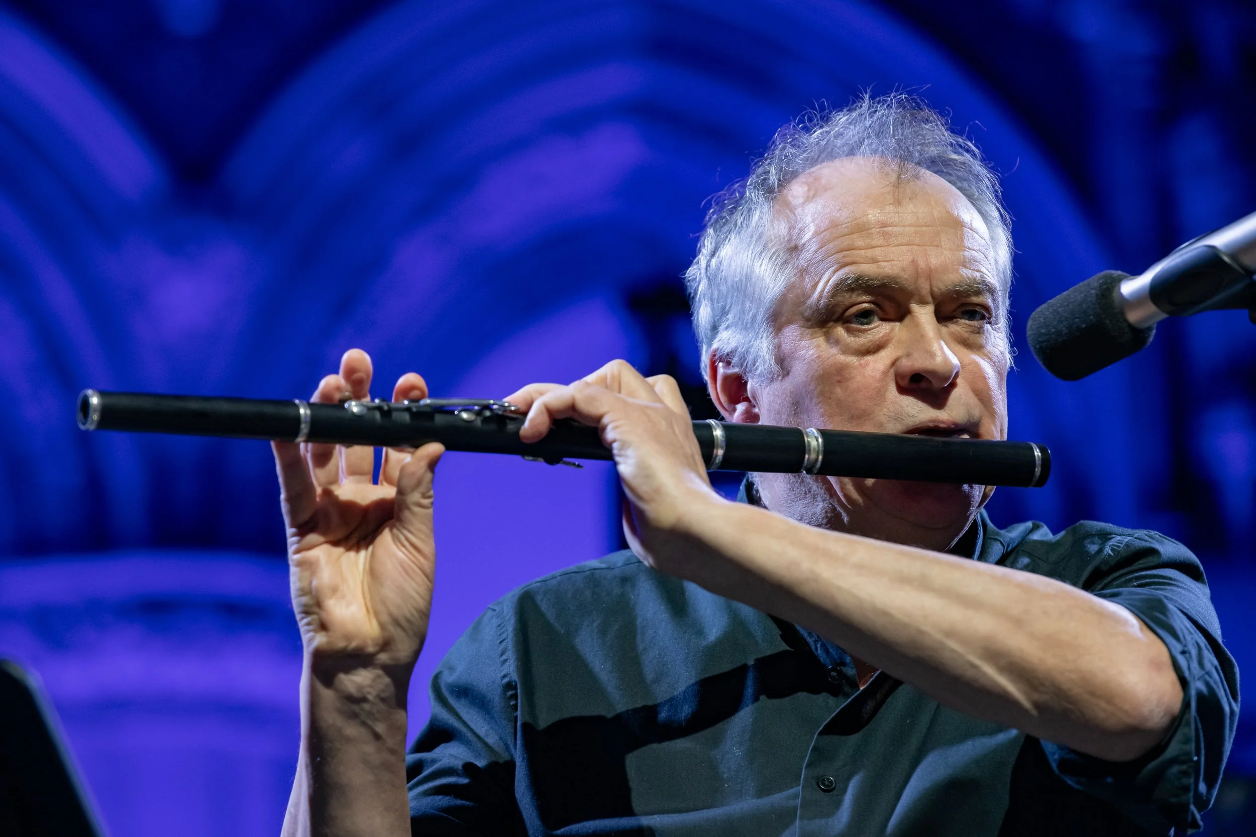 An older man with gray hair playing a black flute on stage, with a microphone nearby and a blurred blue background.