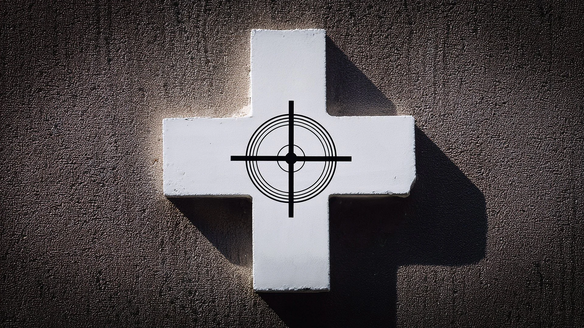 A white stone cross mounted on a textured concrete wall, a dramatic shadow cast behind it, with a black rifle crosshair symbol centered precisely over the intersection of the cross.