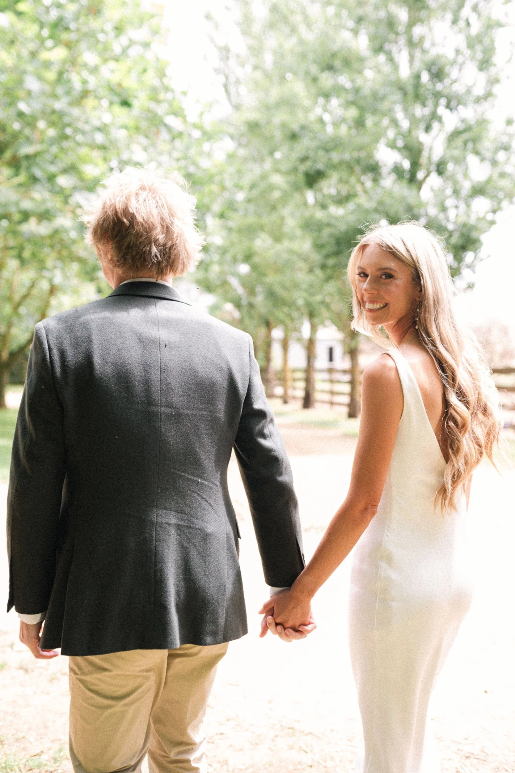 A woman in a white dress holding hands with a man in a dark blazer and beige pants, standing outdoors with trees and a fence in the background.