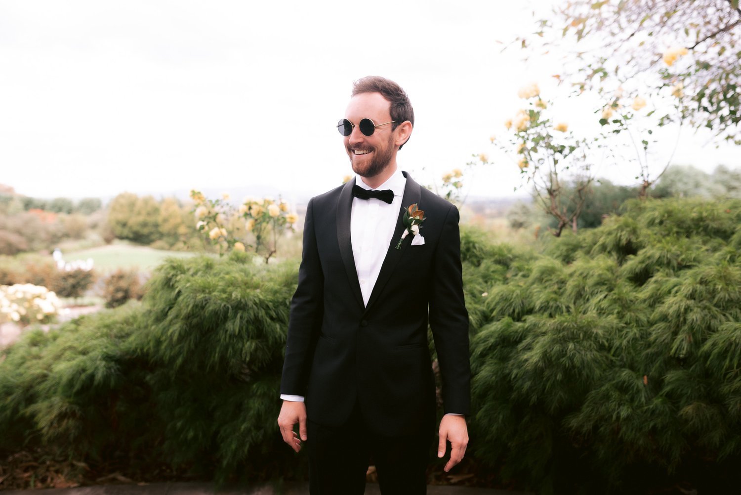 Man in black tuxedo with bow tie and sunglasses smiling, standing in front of lush greenery and yellow flowers.