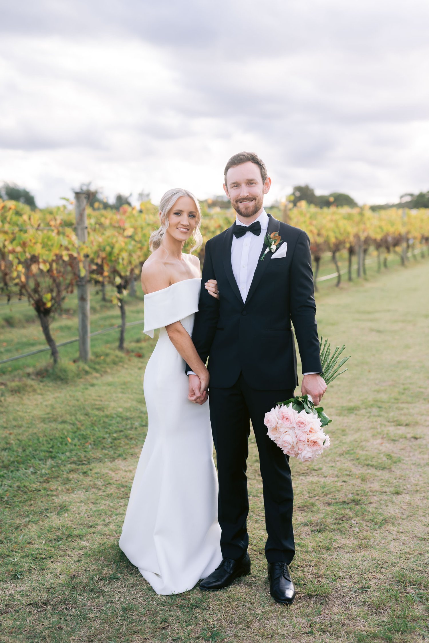Bride and groom standing in a vineyard, bride in white dress, groom in black tuxedo holding bouquet.