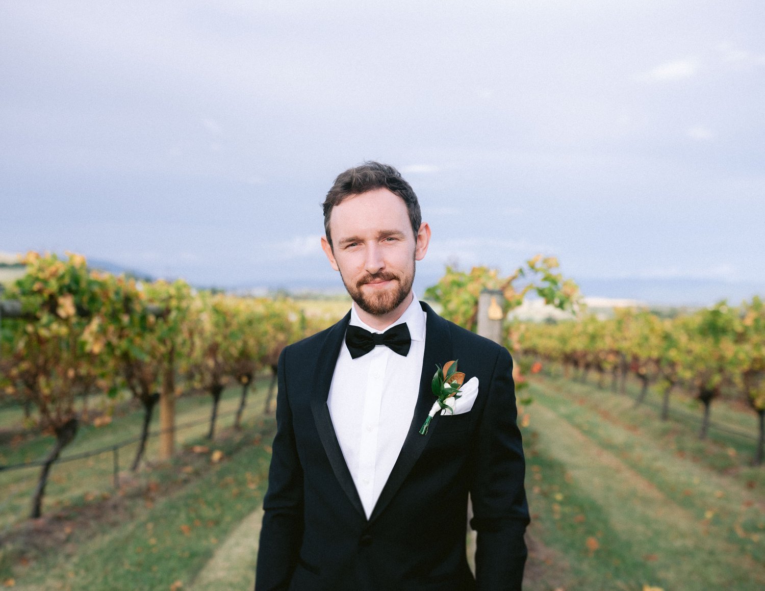 Man in tuxedo with bow tie, standing in vineyard, smiling