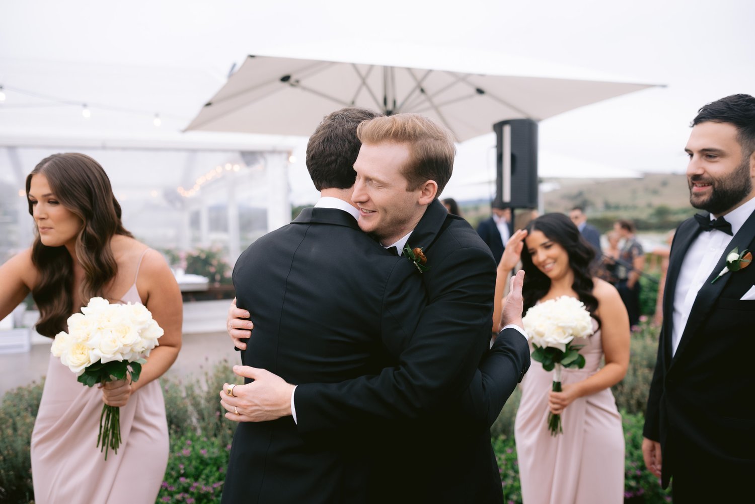 Wedding party celebrating outdoors with people hugging and holding white flower bouquets.