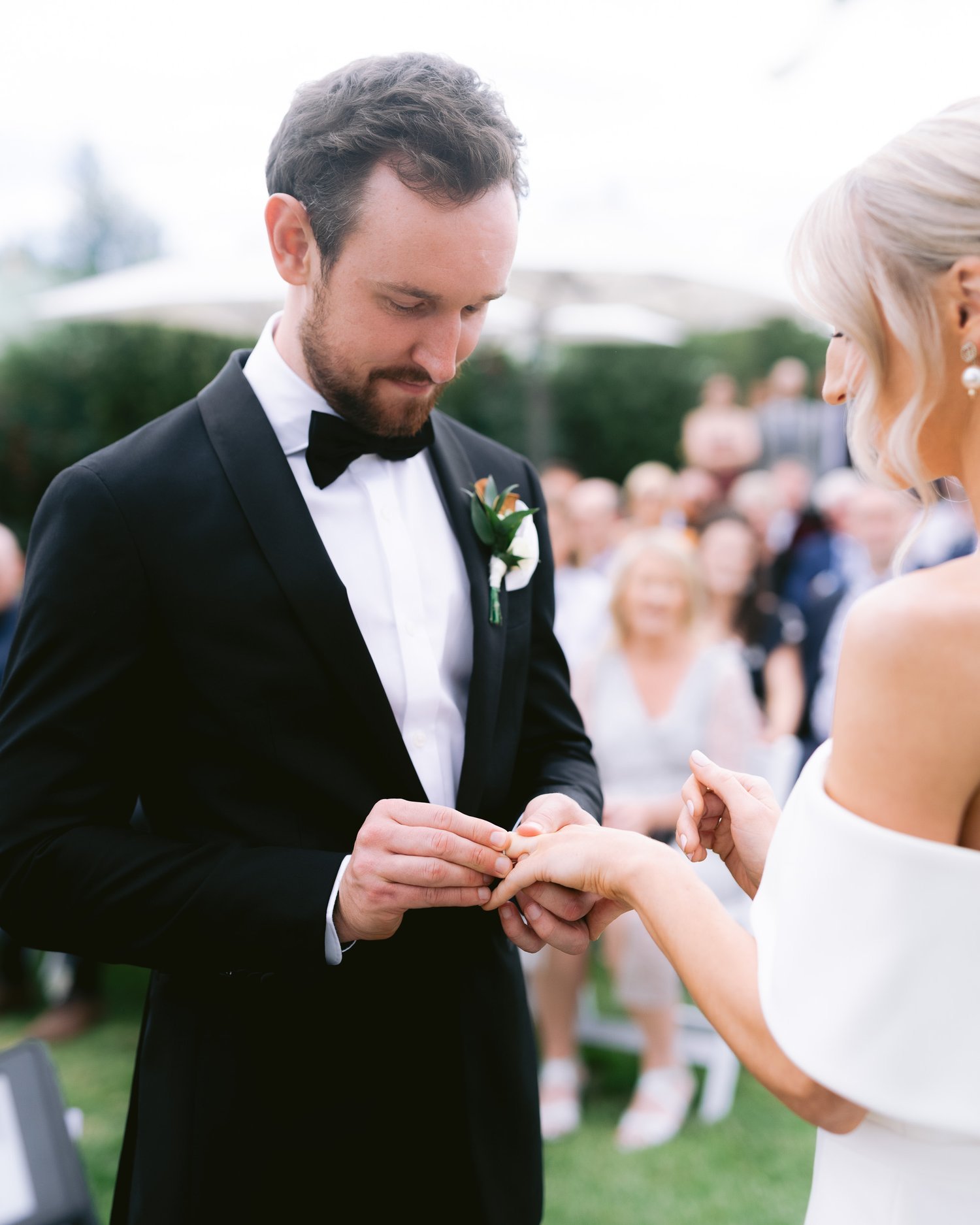 A groom in a black tuxedo placing a wedding ring on the bride's finger during an outdoor ceremony.