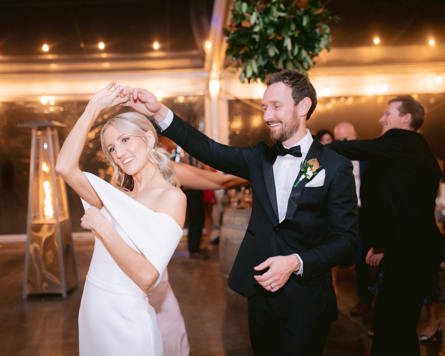 Bride and groom dancing at a wedding reception, bride in a white gown, groom in a black suit with bow tie, surrounded by blurred guests and string lights.