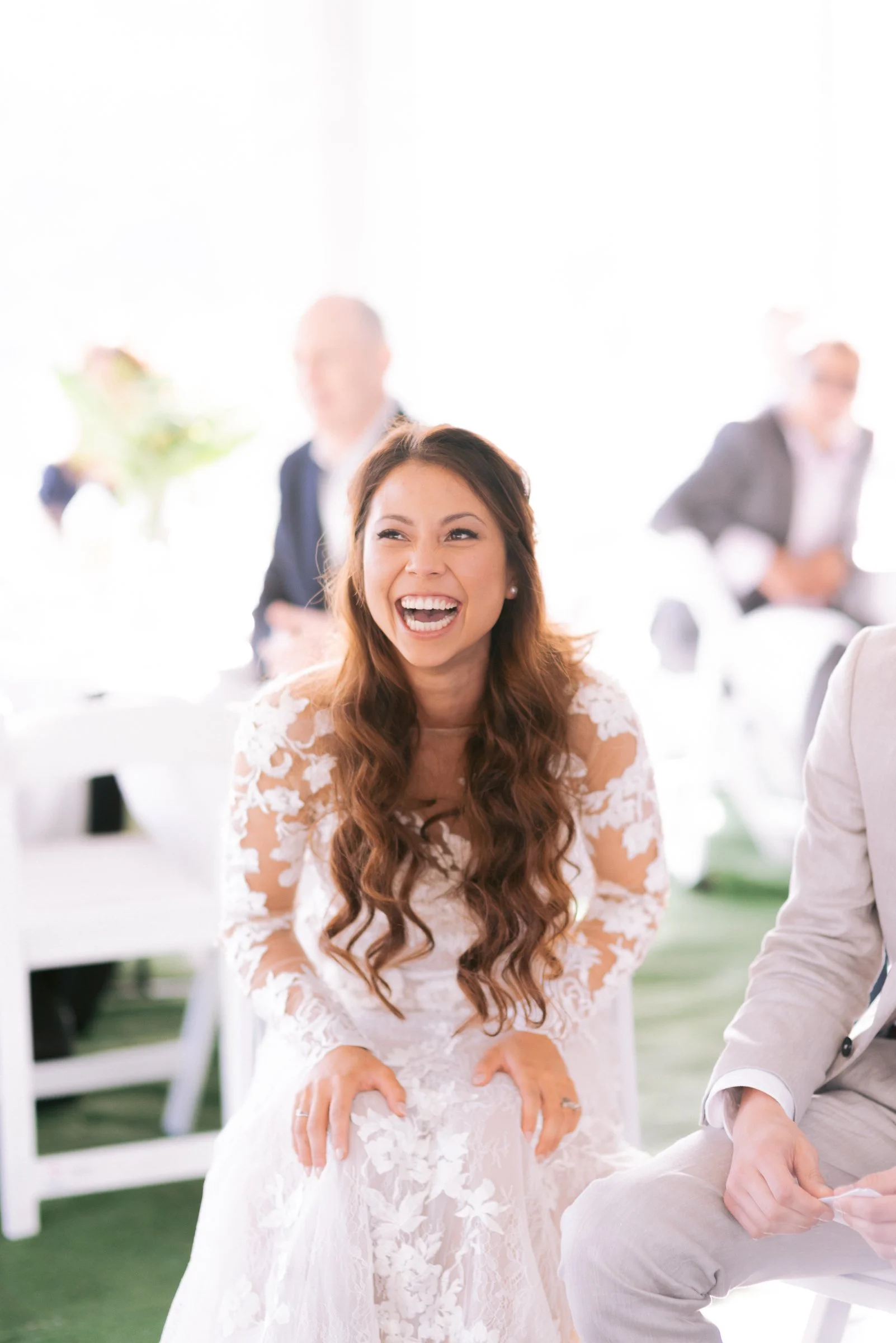 Bride in lace wedding dress smiling and sitting indoors, with guests in the background.