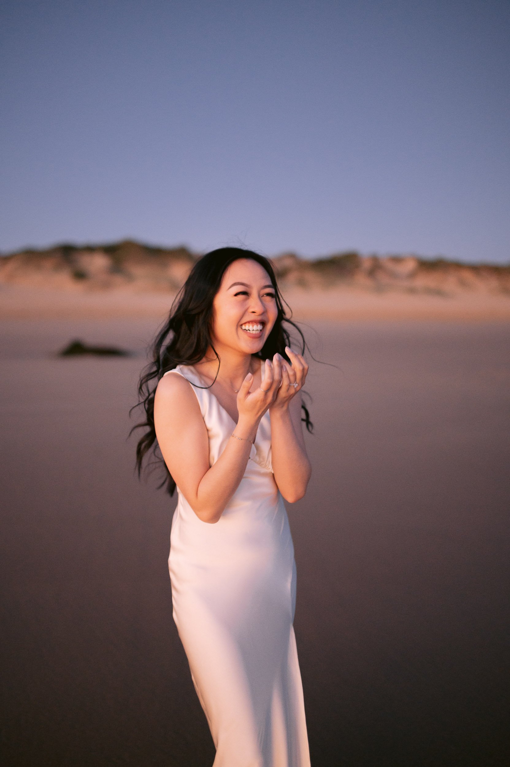 A woman in a white dress laughing and standing on a sandy beach at sunset.