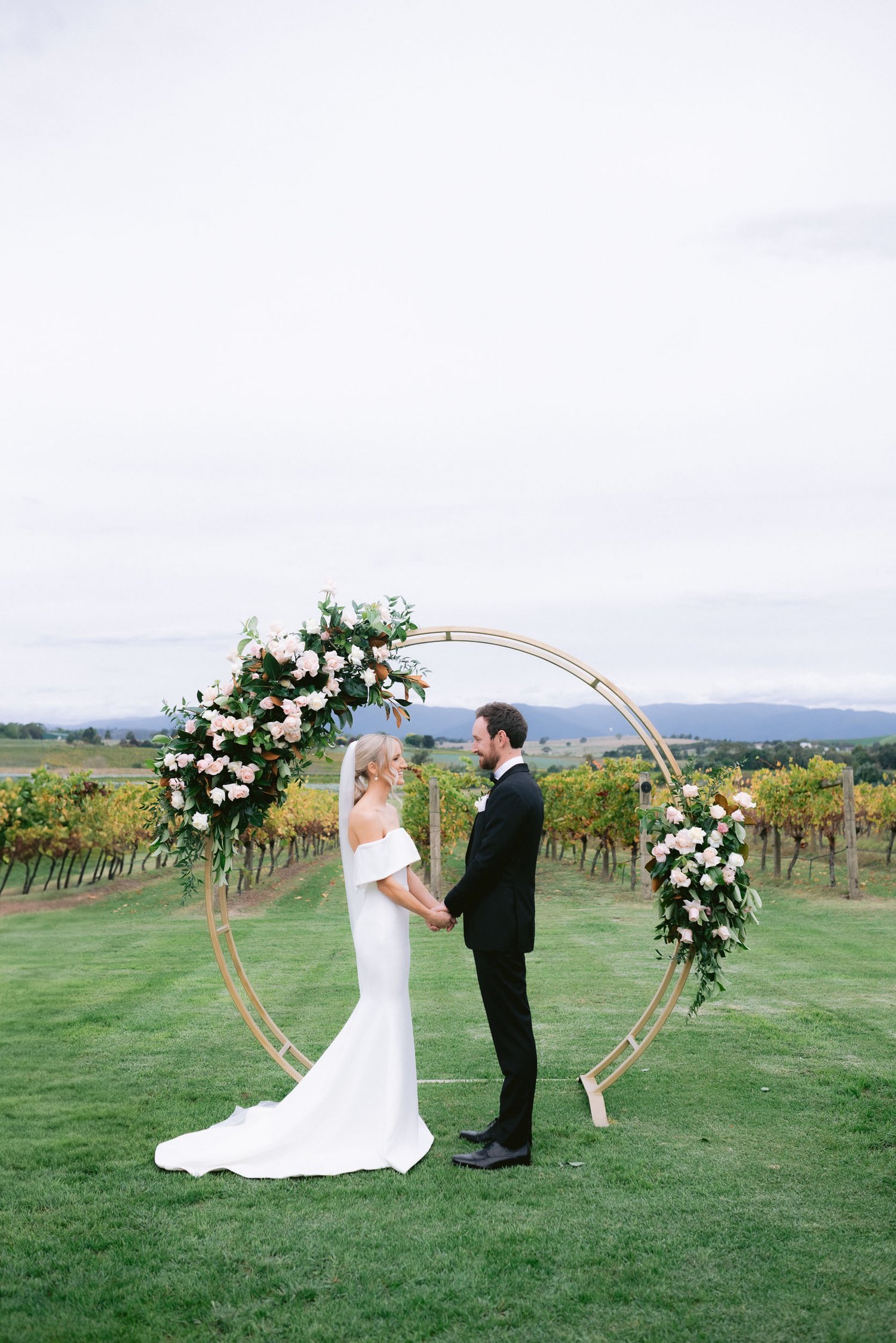 Bride in white gown and groom in black suit holding hands, standing in front of floral circular arch in a vineyard setting.