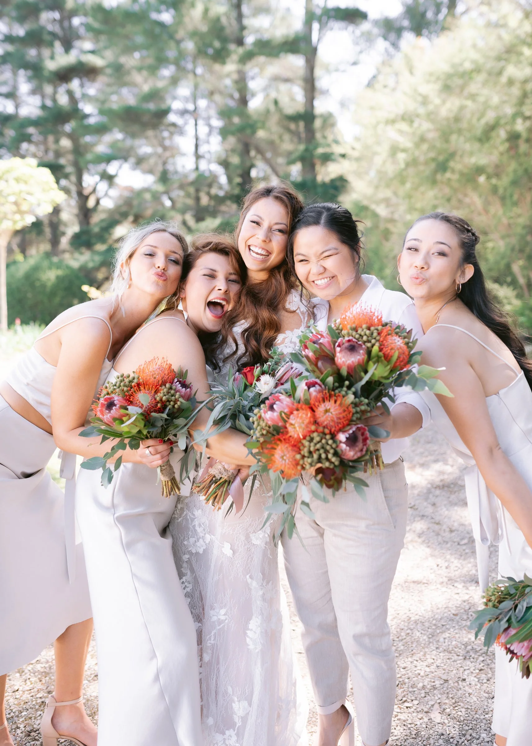 Group of five women smiling and posing with flowers, outdoors in a bright, forested setting.