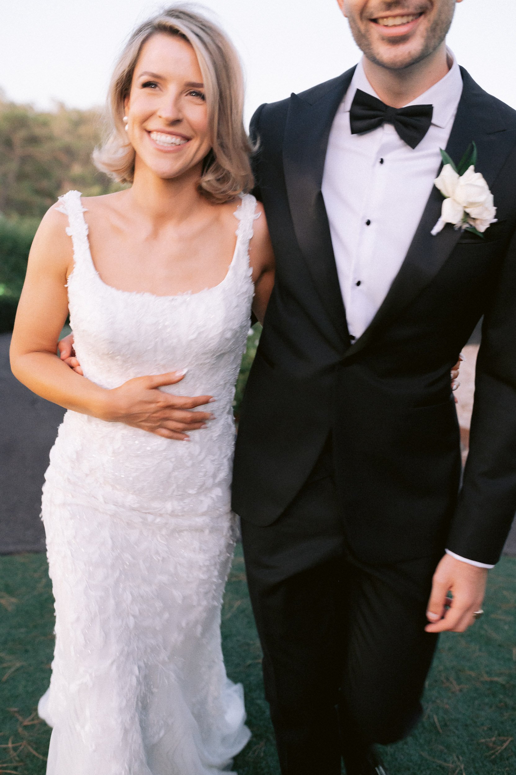 A happy bride in a white wedding dress and a smiling groom in a black tuxedo walk outside on a wedding day.