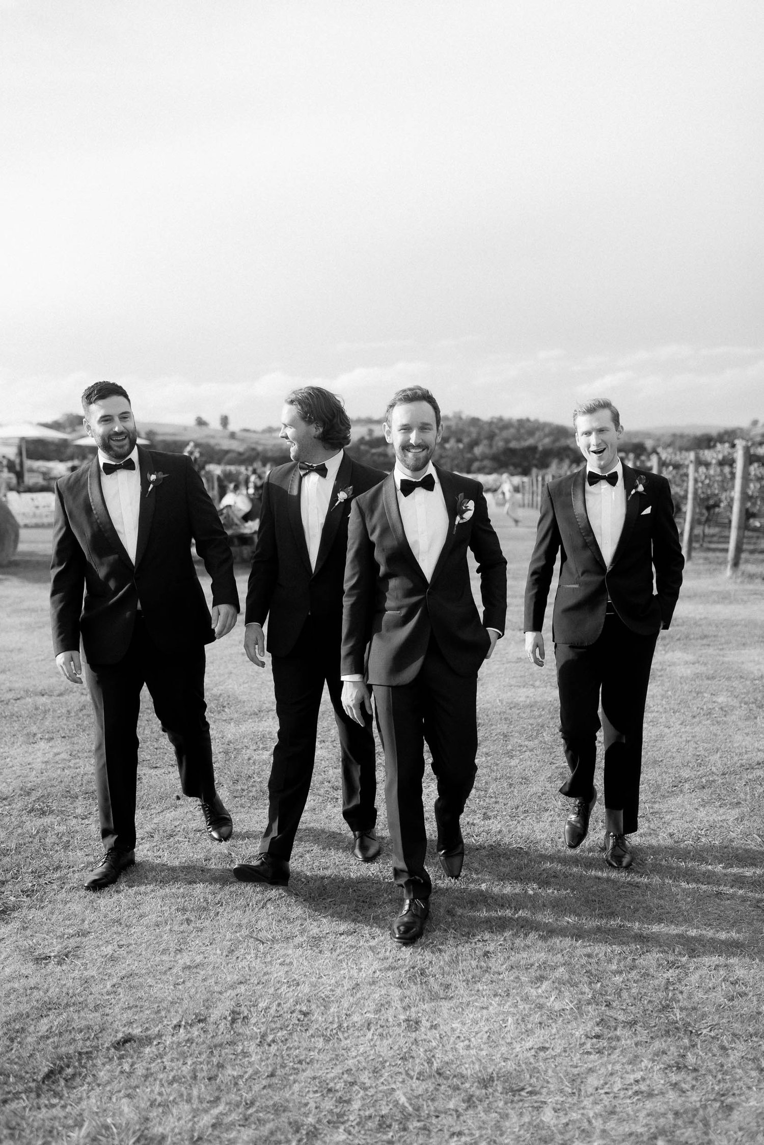 Four men in tuxedos walking outdoors on grass, smiling and chatting, in a black and white photo.