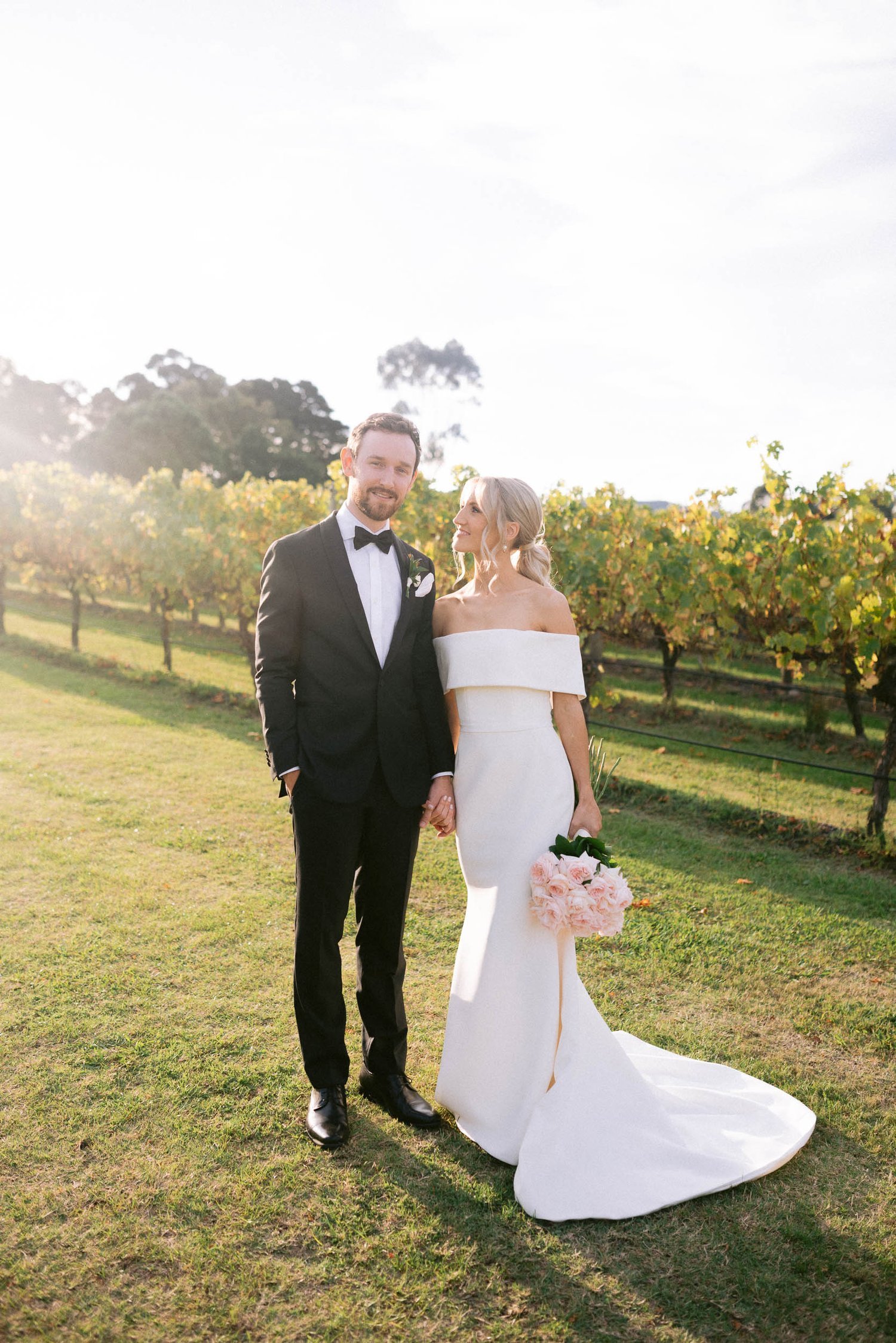 Bride and groom holding hands in a vineyard, bride in white dress with bouquet, groom in black suit and bow tie.