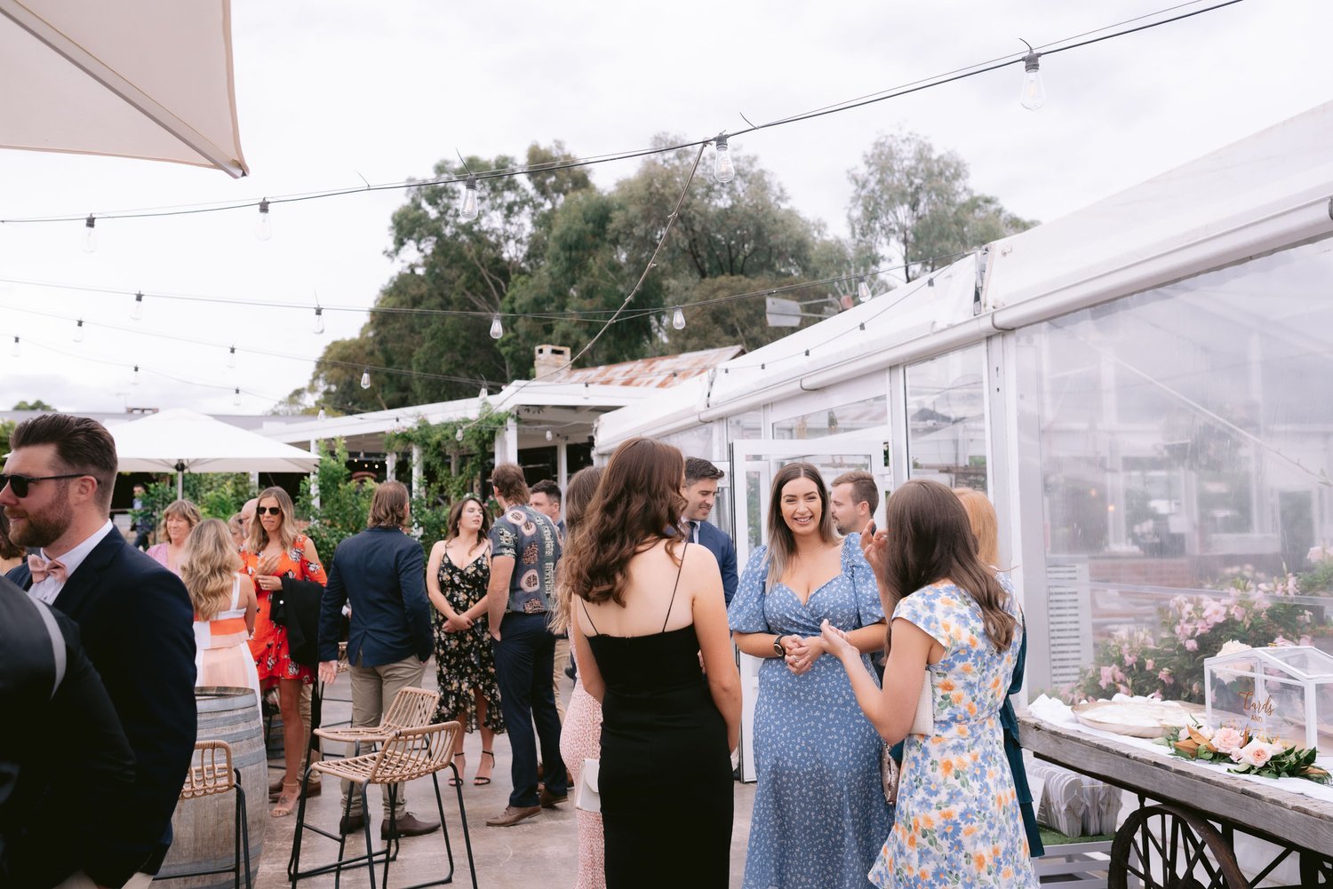 Outdoor event with people in formal attire, string lights overhead, and a tented area. Guests are socializing near tables and chairs.