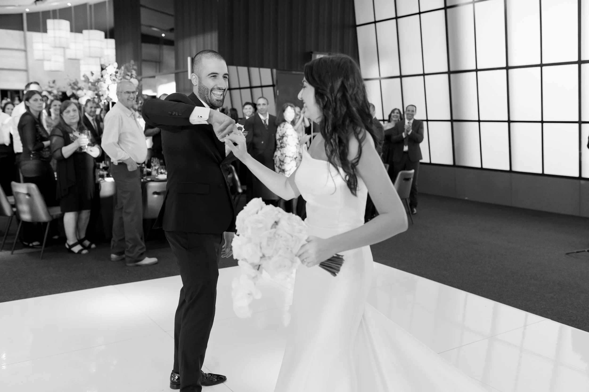 A couple gets married, the groom smiles and holds the bride's hand, surrounded by guests in an indoor wedding venue.