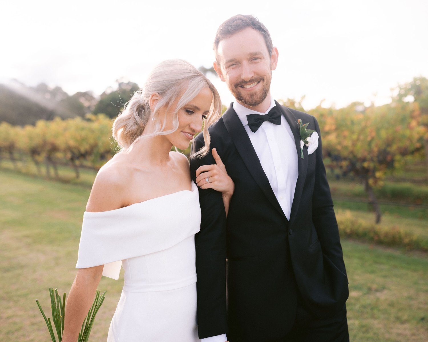 Bride and groom walking in a vineyard, wearing formal wedding attire.
