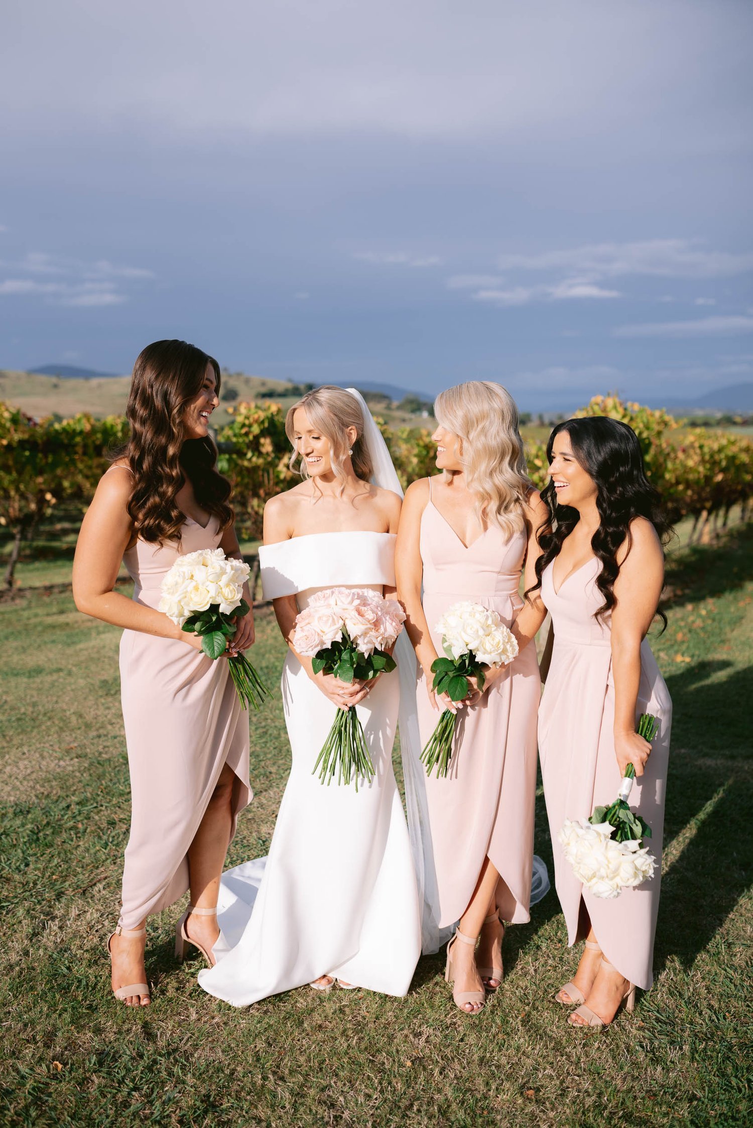 Bride and three bridesmaids in blush dresses holding bouquets, standing outdoors on a sunny day.