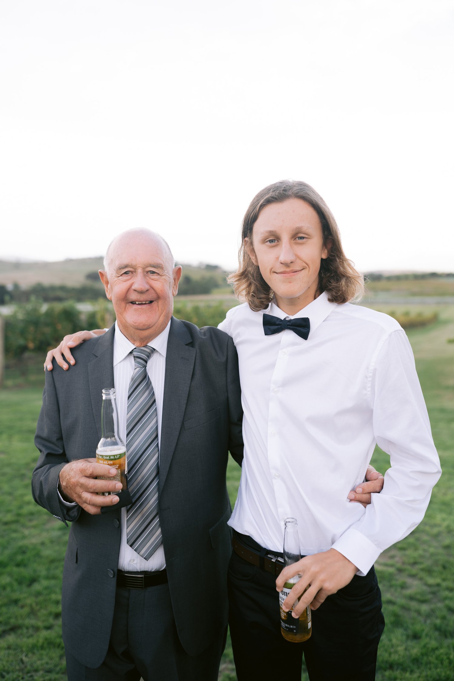 Two people posing on a grassy field, one wearing a suit holding a beer, the other in a white shirt with a bow tie holding a beer.
