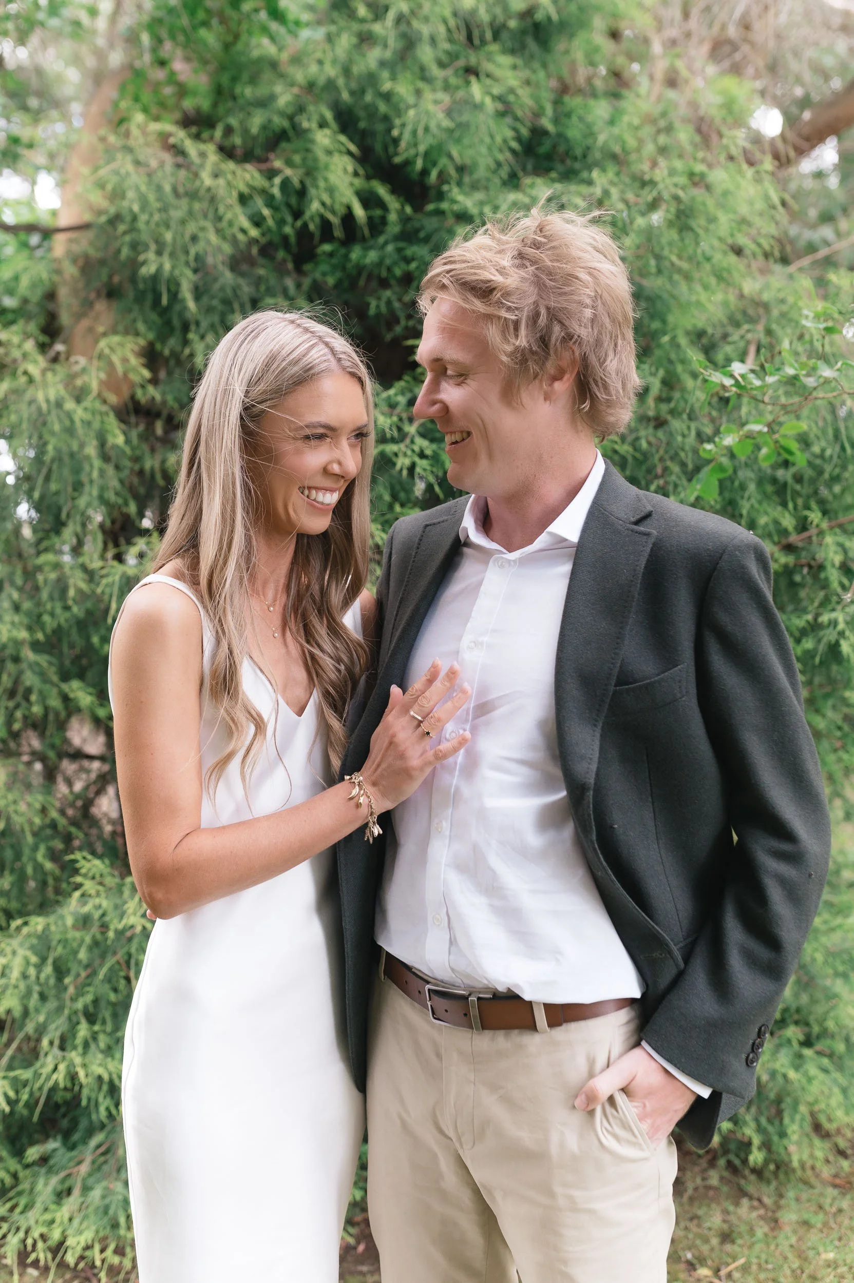 A smiling woman in a white dress touching a man in a white shirt and dark blazer, outdoors with green foliage in the background.