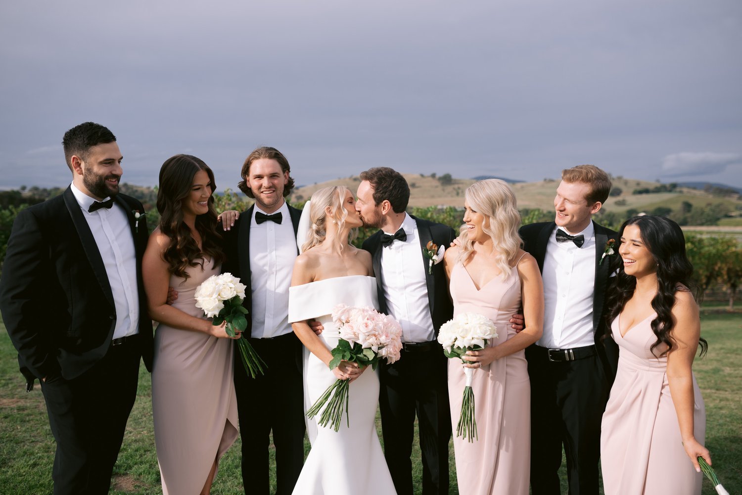 A wedding party posing outdoors, with bridesmaids in pink dresses and groomsmen in black suits. The bride and groom are in the center, embracing and kissing, holding bouquets. The background features a vineyard and hills under a cloudy sky.
