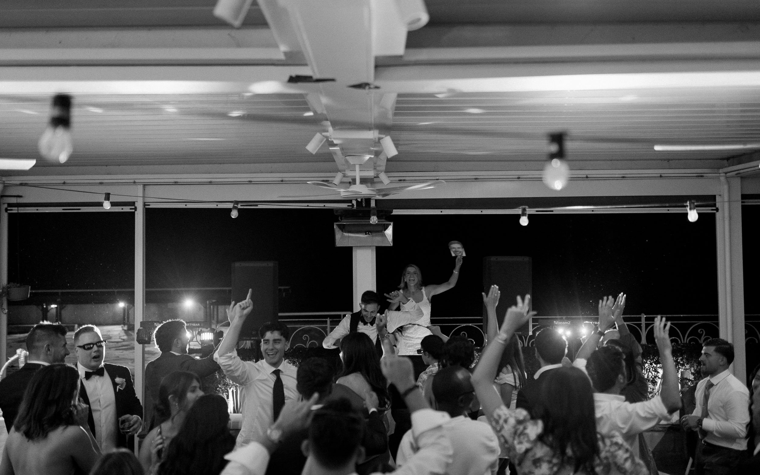 Black and white photo of people dancing and celebrating at a party or wedding reception, with a woman dancing on a table or platform.