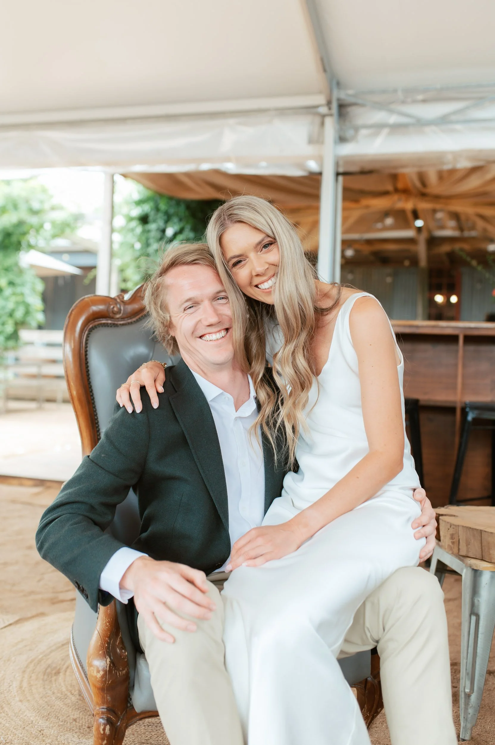 A happy couple sitting on a vintage armchair inside a tent, smiling and embracing at an outdoor event.