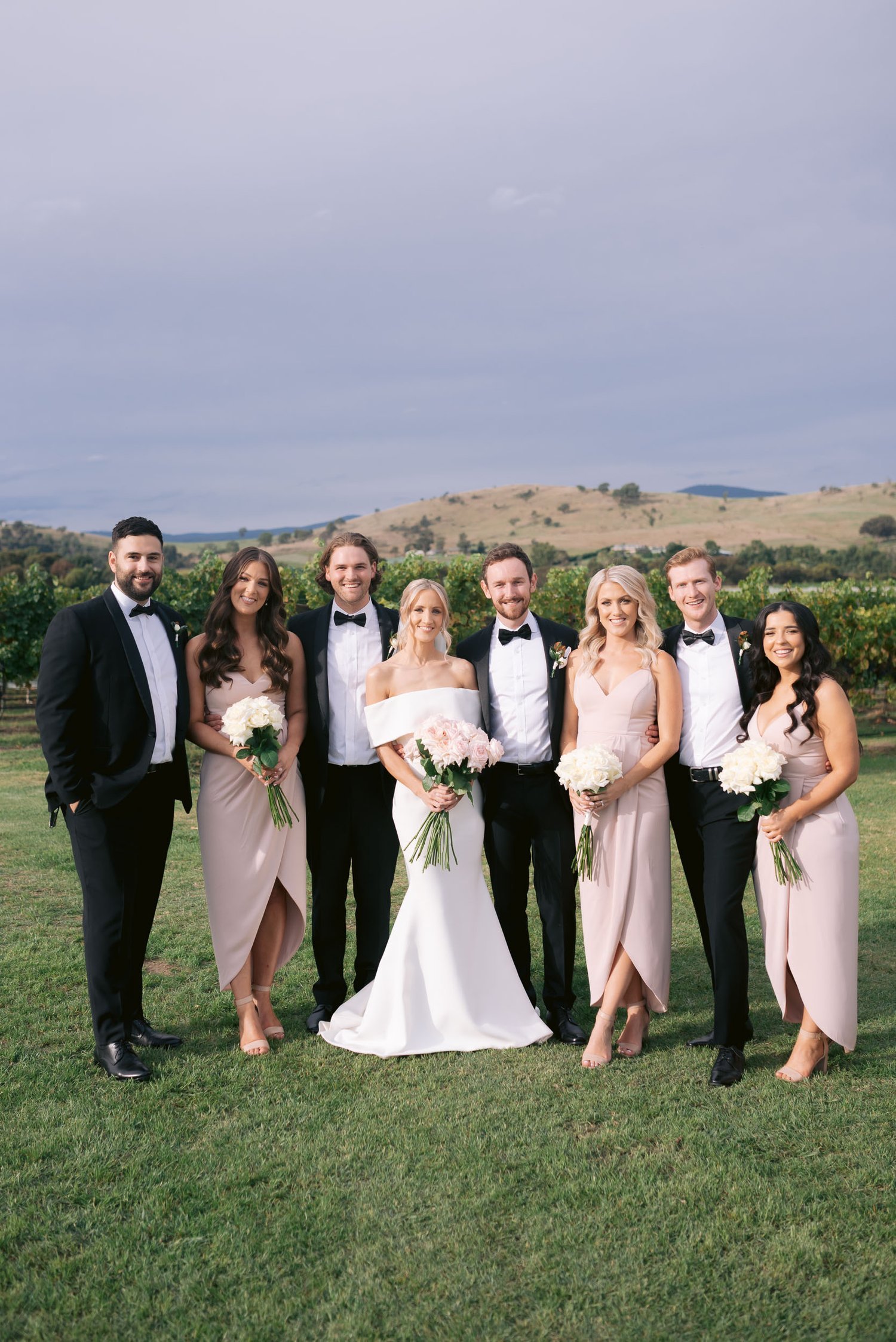 A group of seven people dressed in formal wedding attire posing outdoors on a grassy field with a scenic hillside background. The bridal party consists of women in dresses holding bouquets and men in tuxedos.